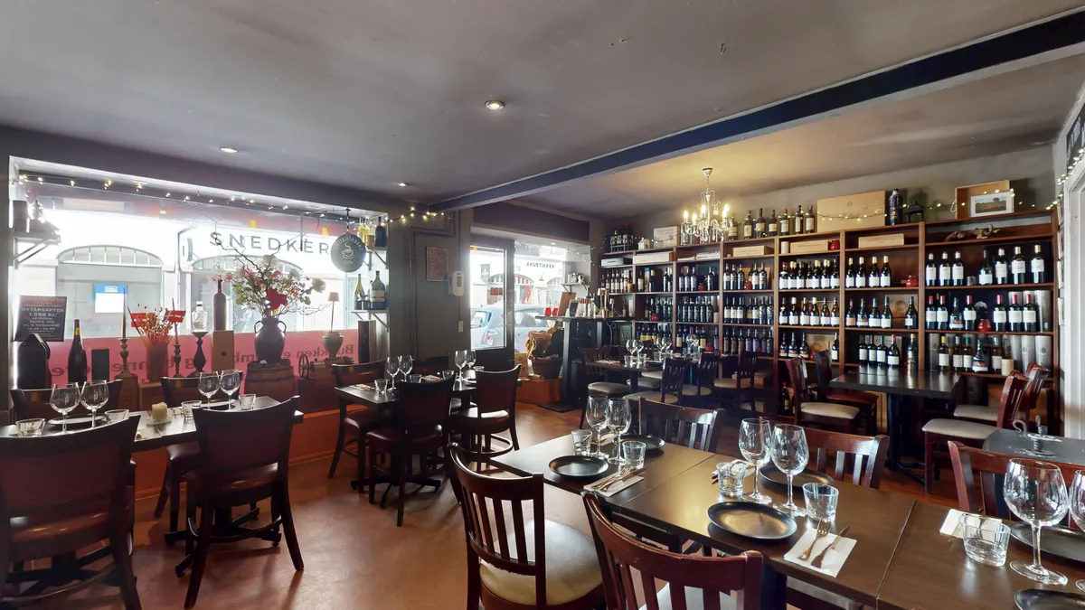 En restaurant med vinflasker på hylder og borde med stole rundt omkring og et gulv med lyse lamper og et vinglas og en vase med blomster