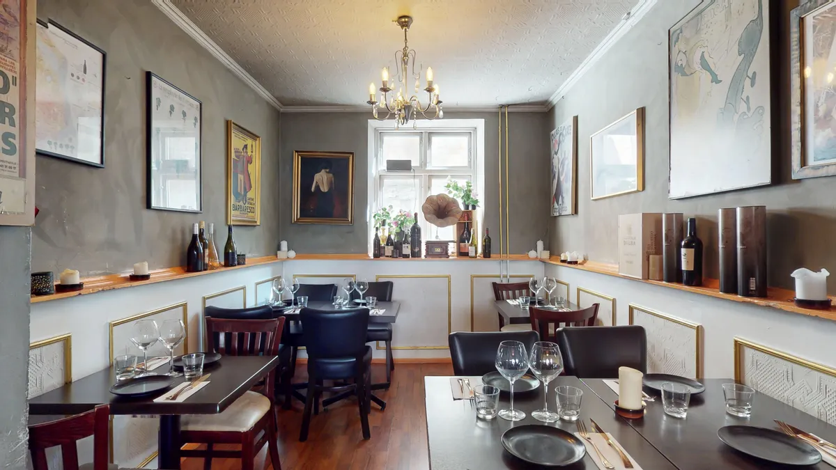 Værelse med borde og stole, vinflasker, krystalliskroner, lysestager og malerier i lyse grå og guld.