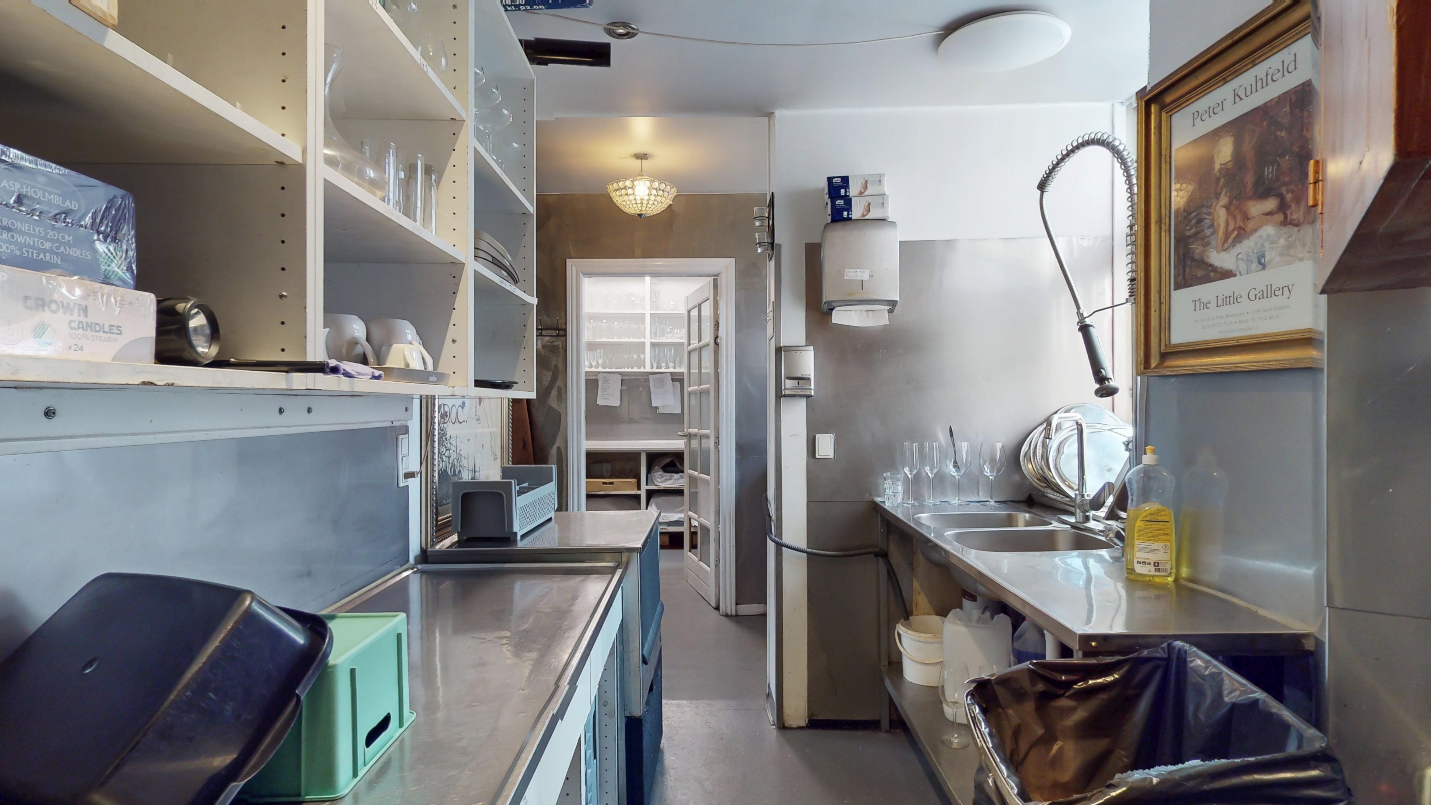 Køkken med højloft og lysekroner, rustfri apparater og ren gulv. Plakat med Peter Kuhfeld og flaske med olie. Hylder med glas og lysekroner.