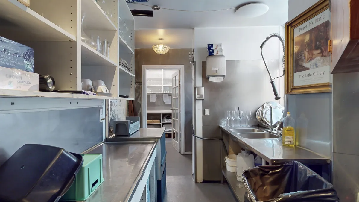 Køkken med højloft og lysekroner, rustfri apparater og ren gulv. Plakat med Peter Kuhfeld og flaske med olie. Hylder med glas og lysekroner.