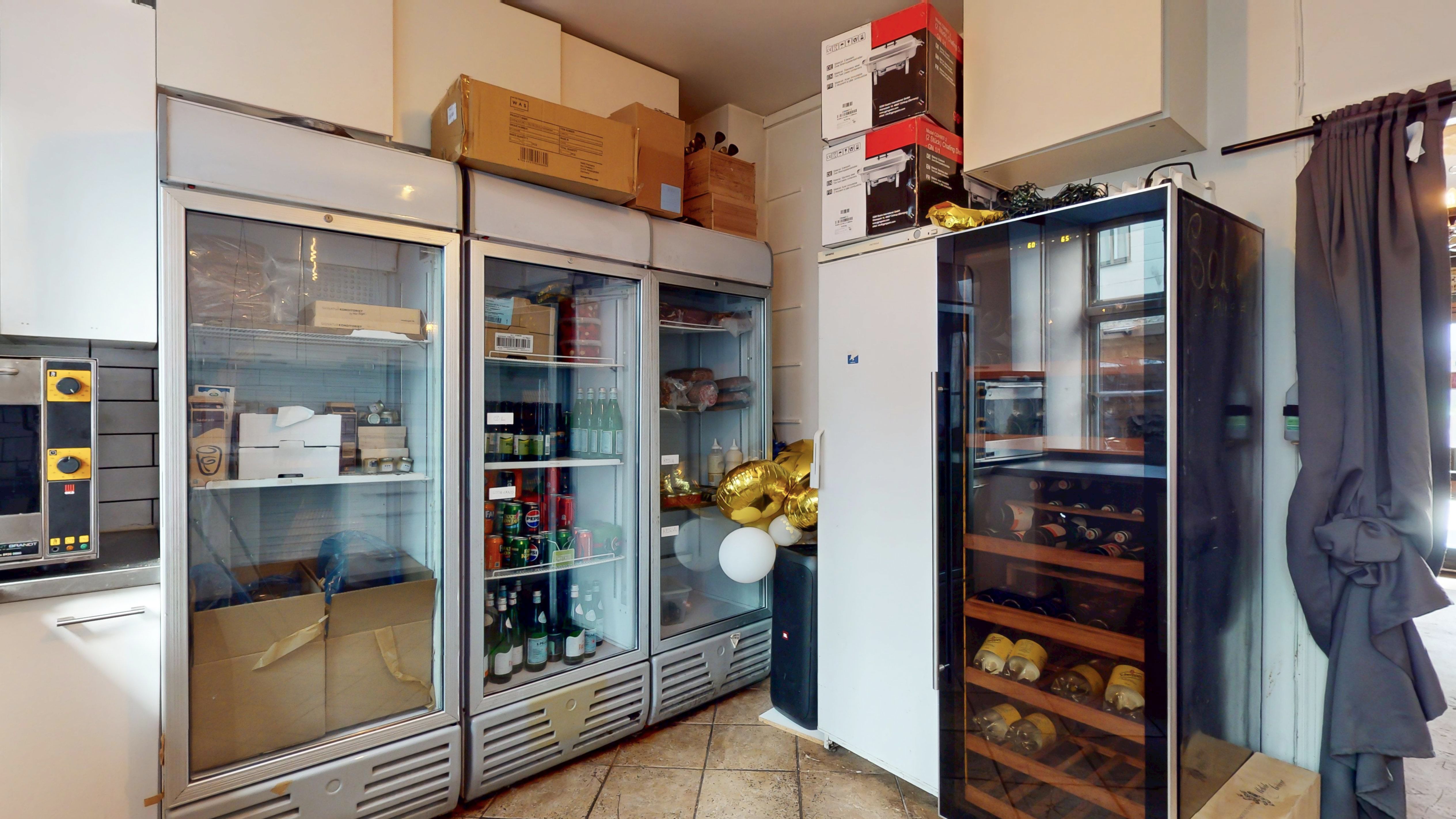 En række køleskabe med drikkevarer og madvarer i et køkken med krydderikasse og flere æskepakker og kasser på hylden