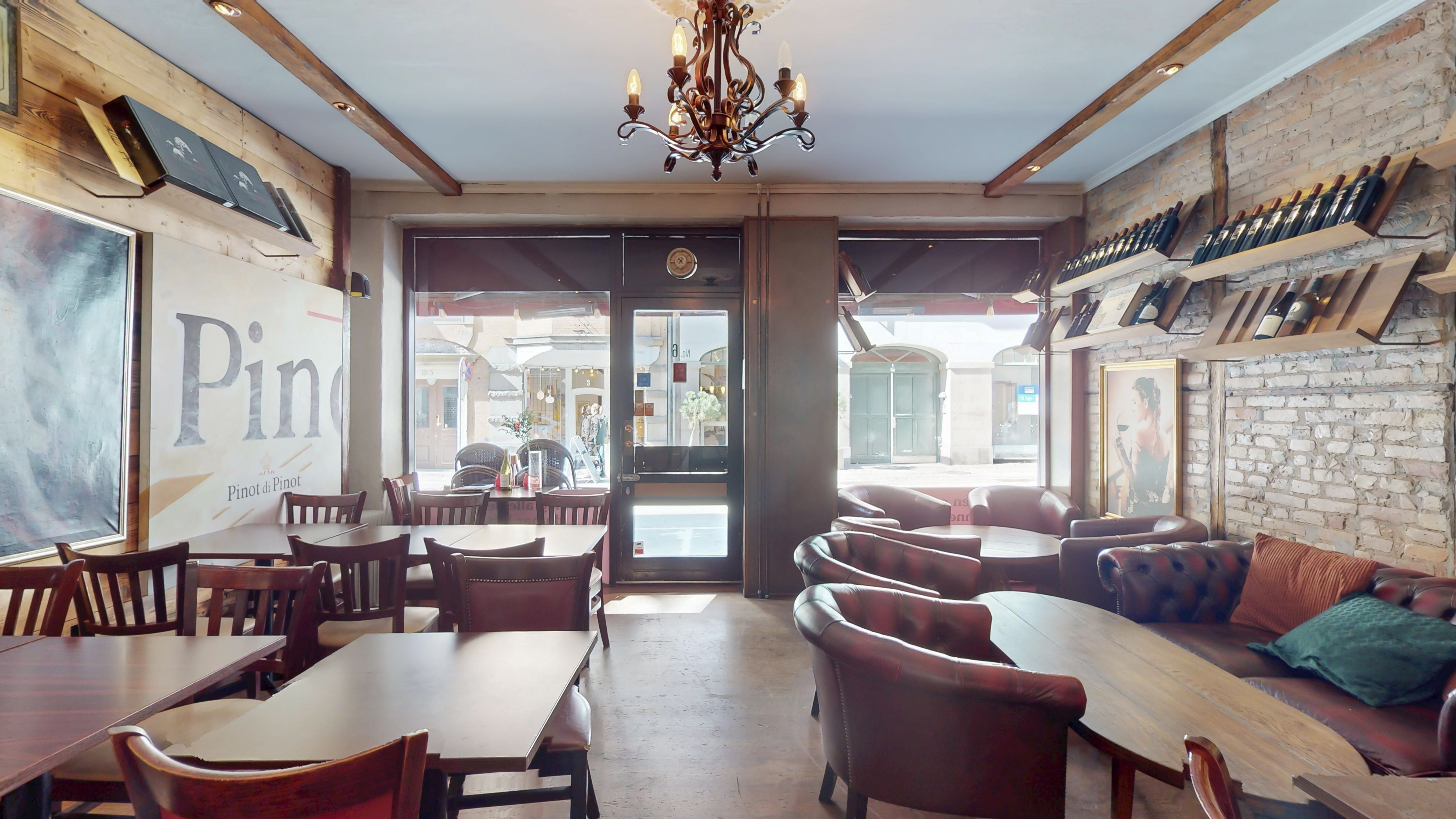 Rustikke spisestue med lyse hjørneborde og læderstole, champagneflasker og et gulv af træ. Lyserød og grøn pude på en lædersofa langs en mur med lyse mursten