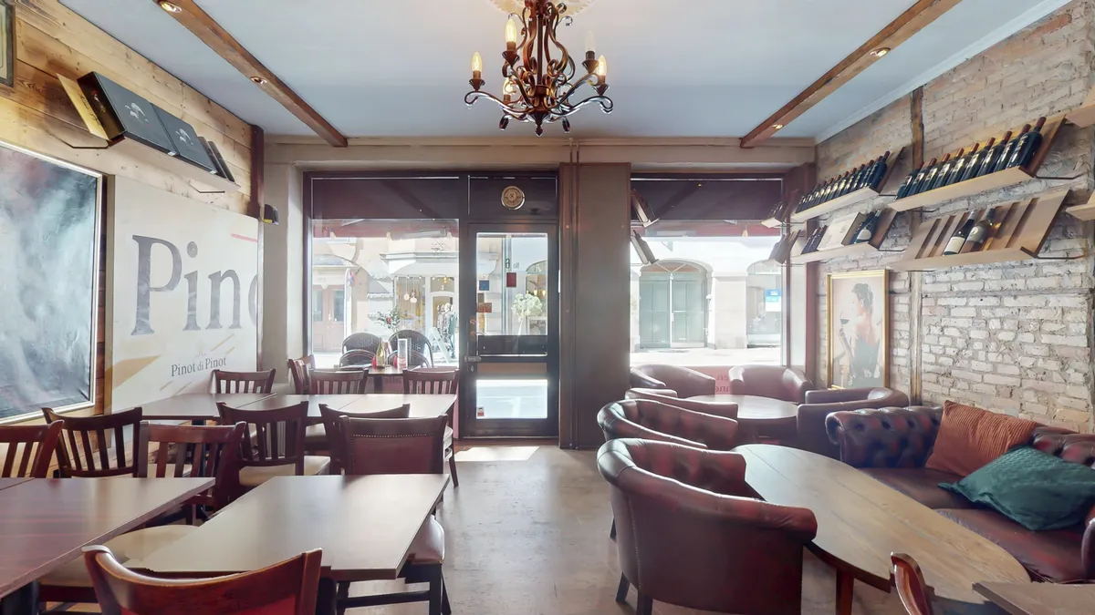 Rustikke spisestue med lyse hjørneborde og læderstole, champagneflasker og et gulv af træ. Lyserød og grøn pude på en lædersofa langs en mur med lyse mursten