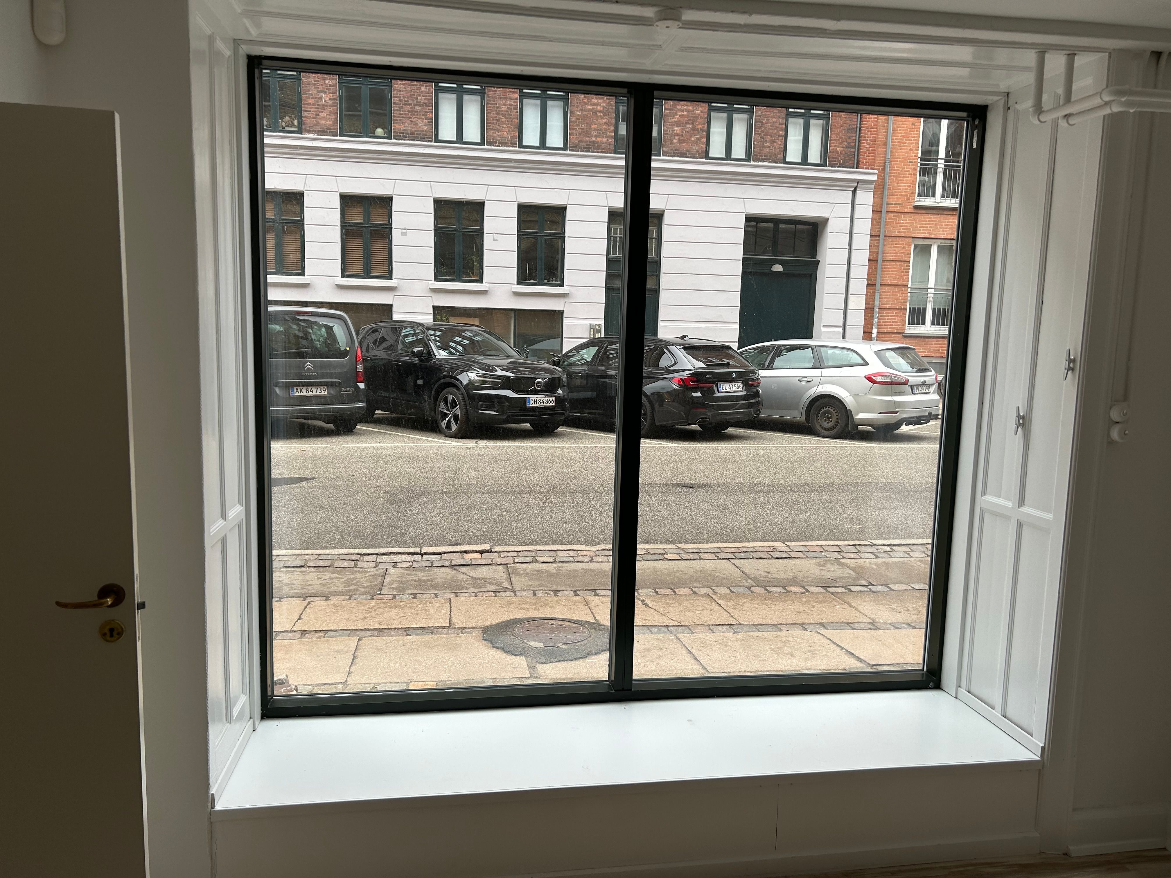 Hvidt, moderne kontorfönster med sort kant, der vender ud mod Ryesgade 72. Først i rækken parkeret er en Citroen og en Volvo. Der er en ny Renault og en Ford efter.