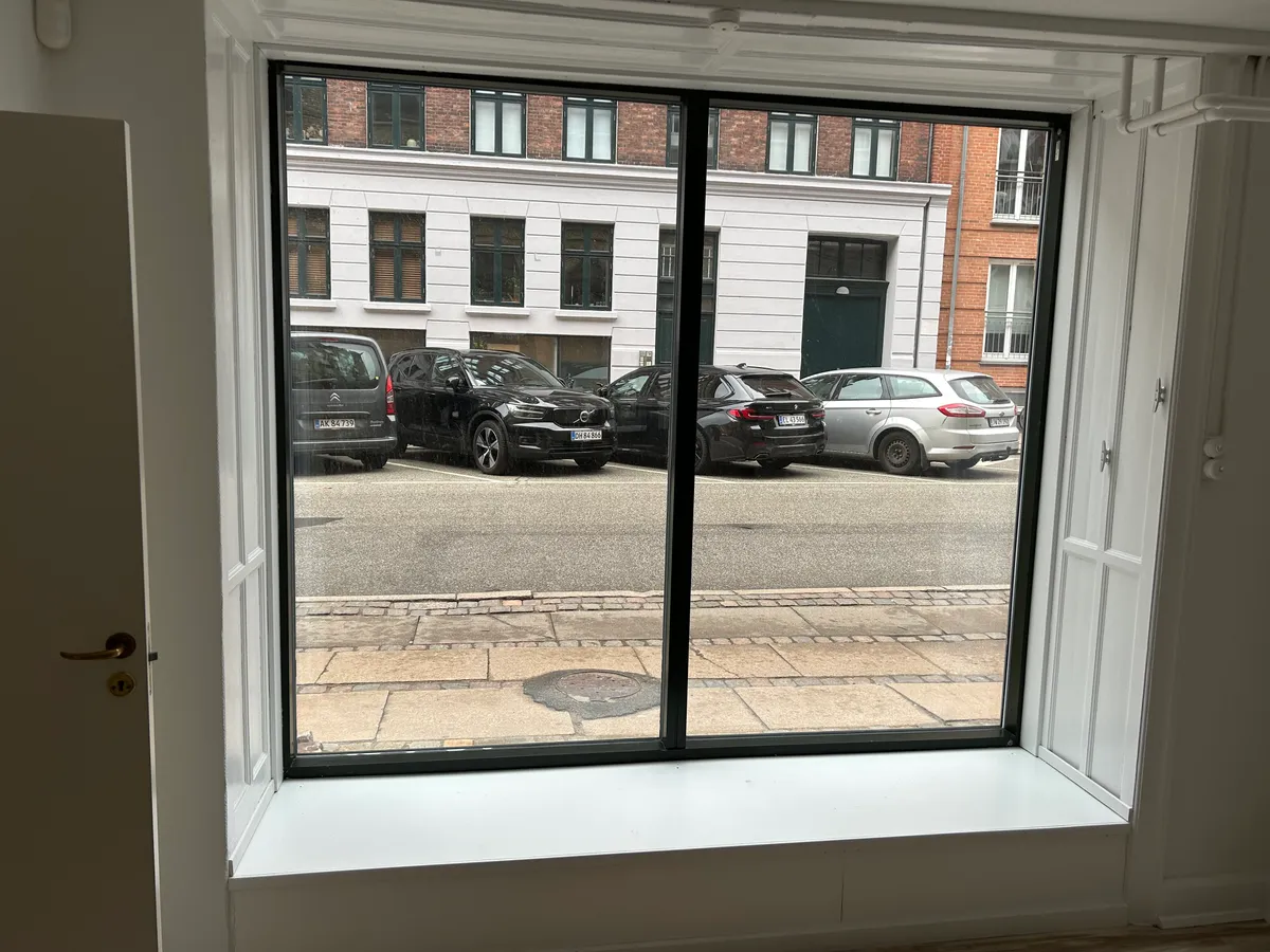 Hvidt, moderne kontorfönster med sort kant, der vender ud mod Ryesgade 72. Først i rækken parkeret er en Citroen og en Volvo. Der er en ny Renault og en Ford efter.