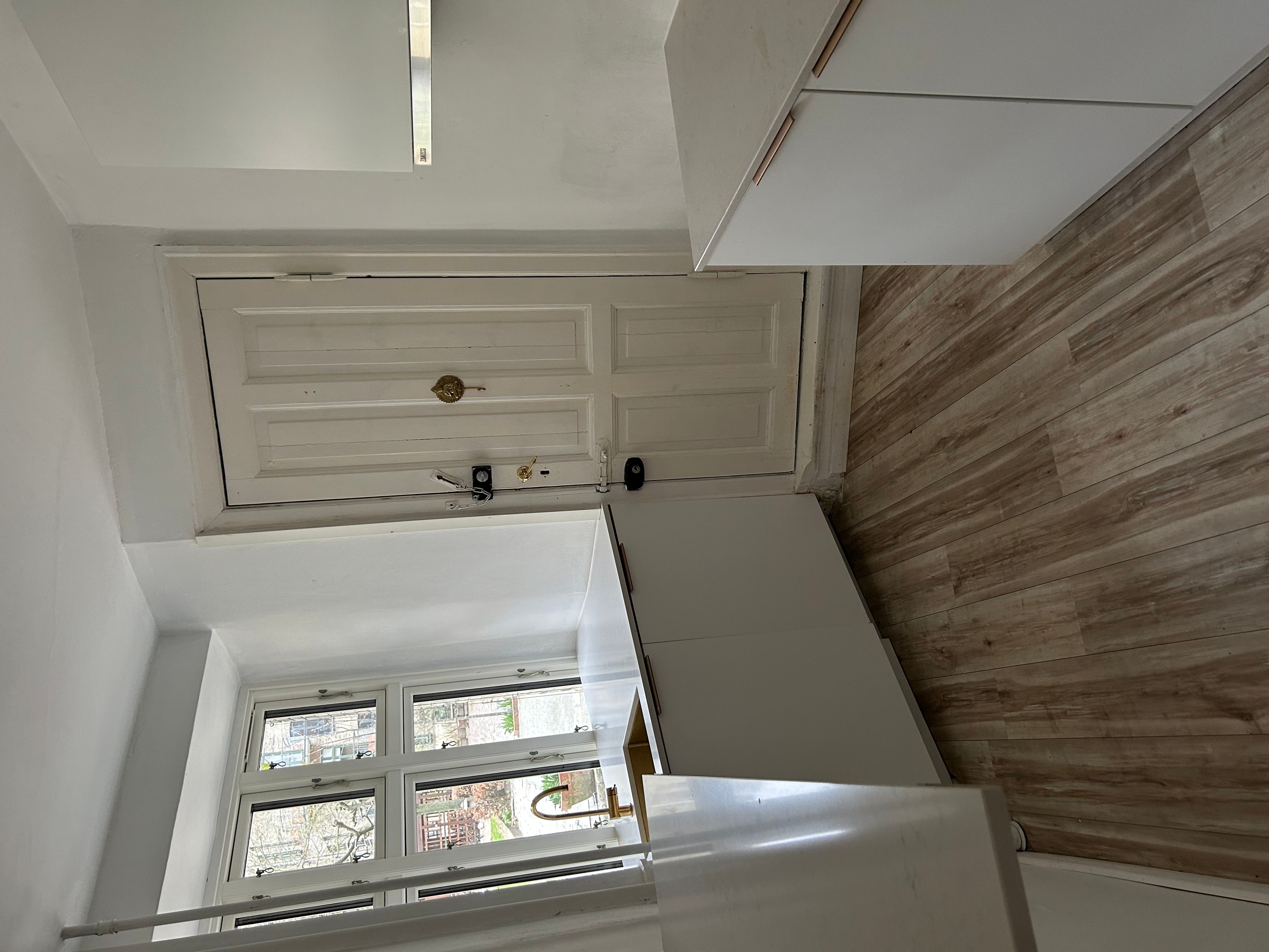 Lukket kontordør i hvidt med guldbeslag. Moderne hvidt køkken med håndvask og vinduer med udsigt til gaden. Væggene er hvide og gulvbelægningen er i lysebrunt træ