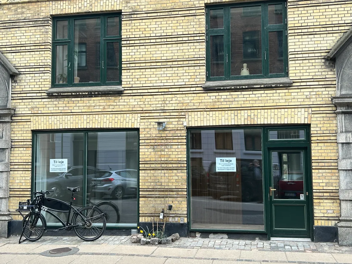 Lejlighed med glasdøre og træindgangsdør med skilte, der siger "Til leje" i forgrunden og en parkeret elektrisk cykel og et reflekteret bilde af biler i baggrunden