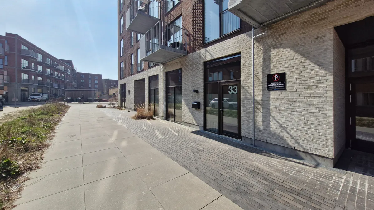 Kontorlokale med store glasruder og metaldøre på Aage Knudsens Strøg 33. Stykkevis udstykket beton og skraldkurve ved siden af. P-skilt med rødt P og forbudsskilt.