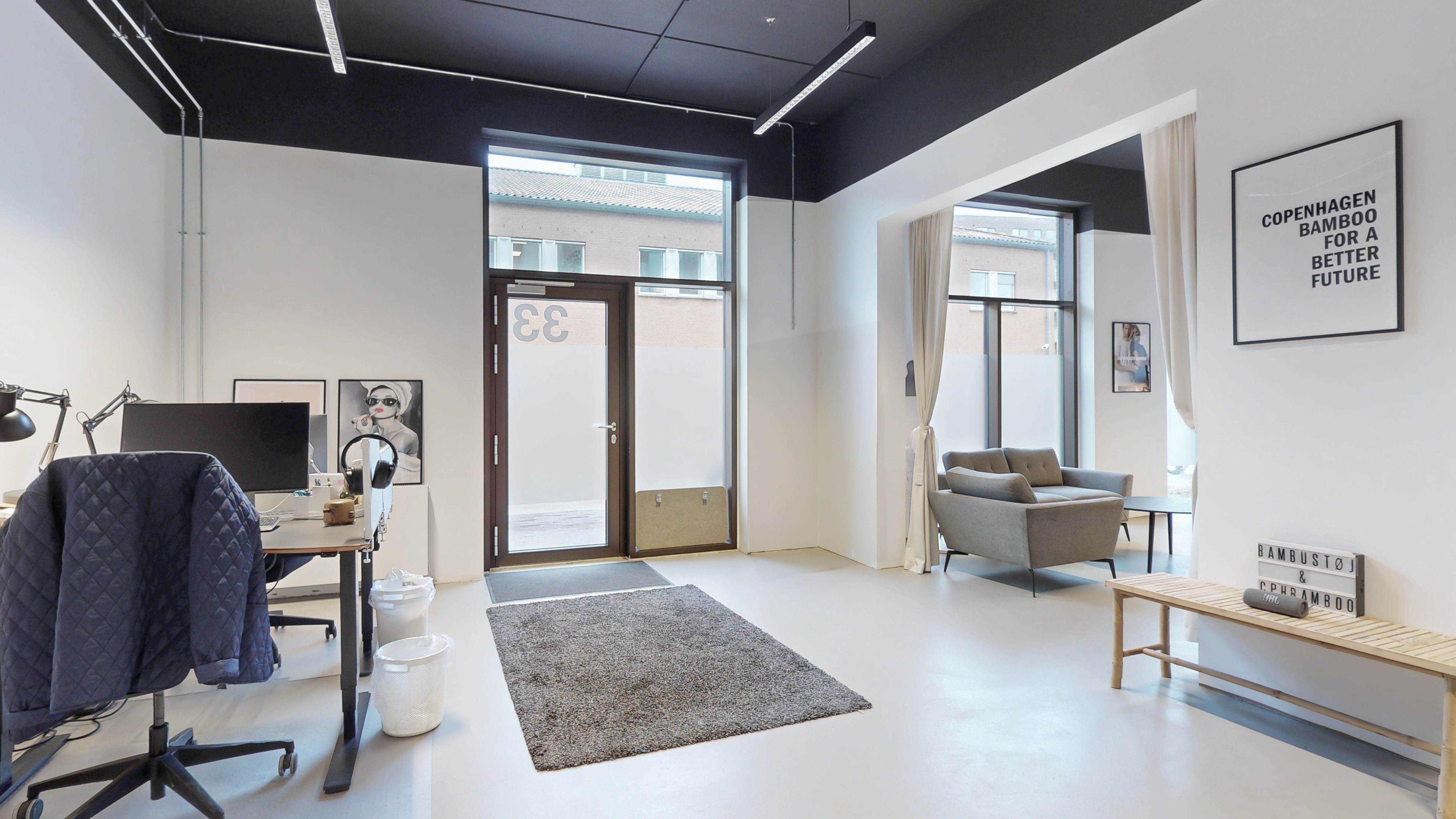 Moderne og lyst kontor med højt loft, hvidt væg og tæppe, med skrivebord, stol og skrivebordslampe samt sovesofa, stolpelampe og tekst på væggen