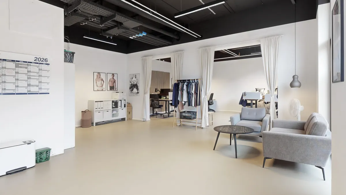 Kontorlokale med hvide vægge, højloft og lyse loftslamper. Et moderne skrivebord med to stole og en stol med tøjvægge. To sofaer og et træbord i højre side.