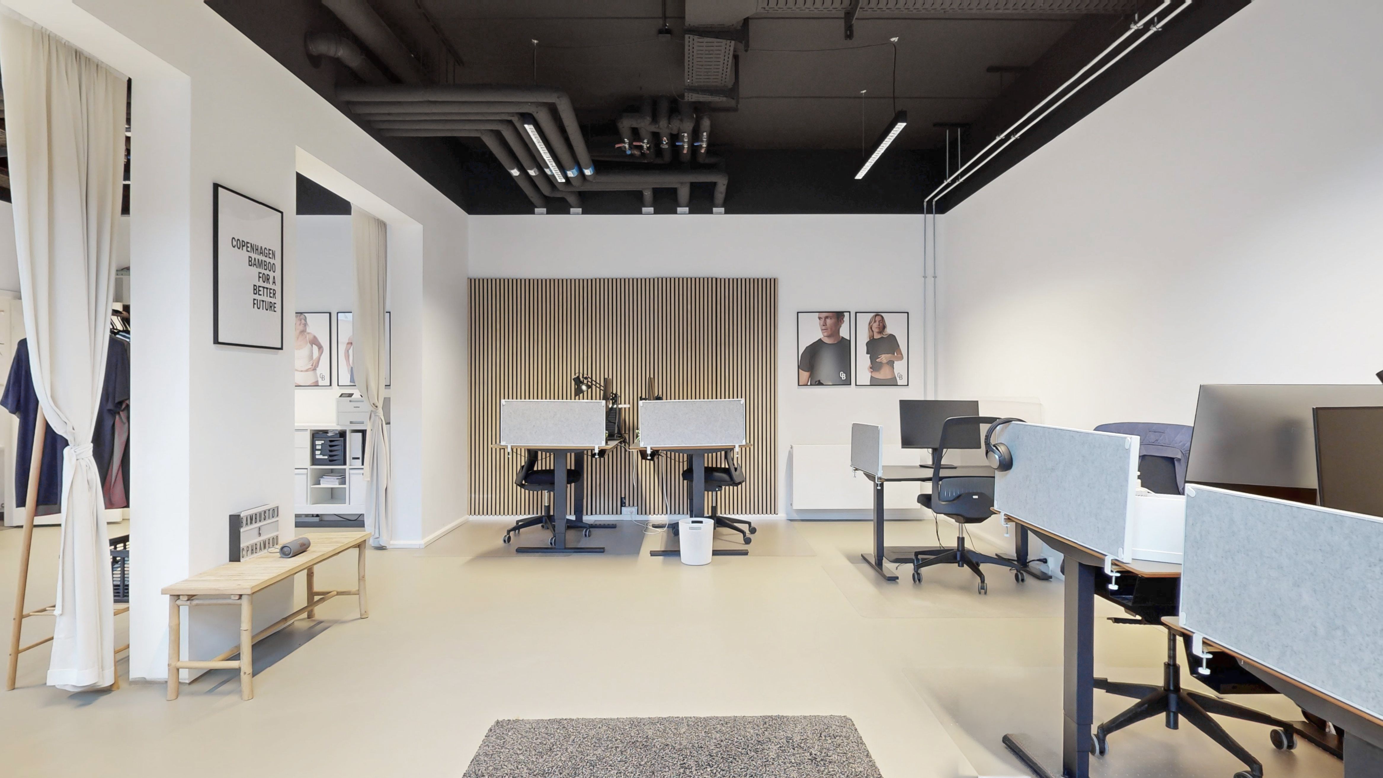 Lyst og moderne kontorlokale med højloft og hvidt vægbeklædning. Kontorlokalet er udstyret med skriveborde, stole, bærbare computere og skrivebordslamper. To billeder hænger på væggen, og der er en hvid bordlampe på skrivebordet.