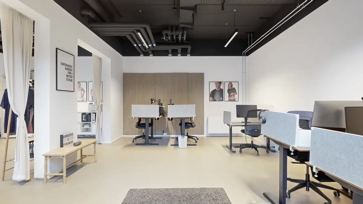 Lyst og moderne kontorlokale med højloft og hvidt vægbeklædning. Kontorlokalet er udstyret med skriveborde, stole, bærbare computere og skrivebordslamper. To billeder hænger på væggen, og der er en hvid bordlampe på skrivebordet.