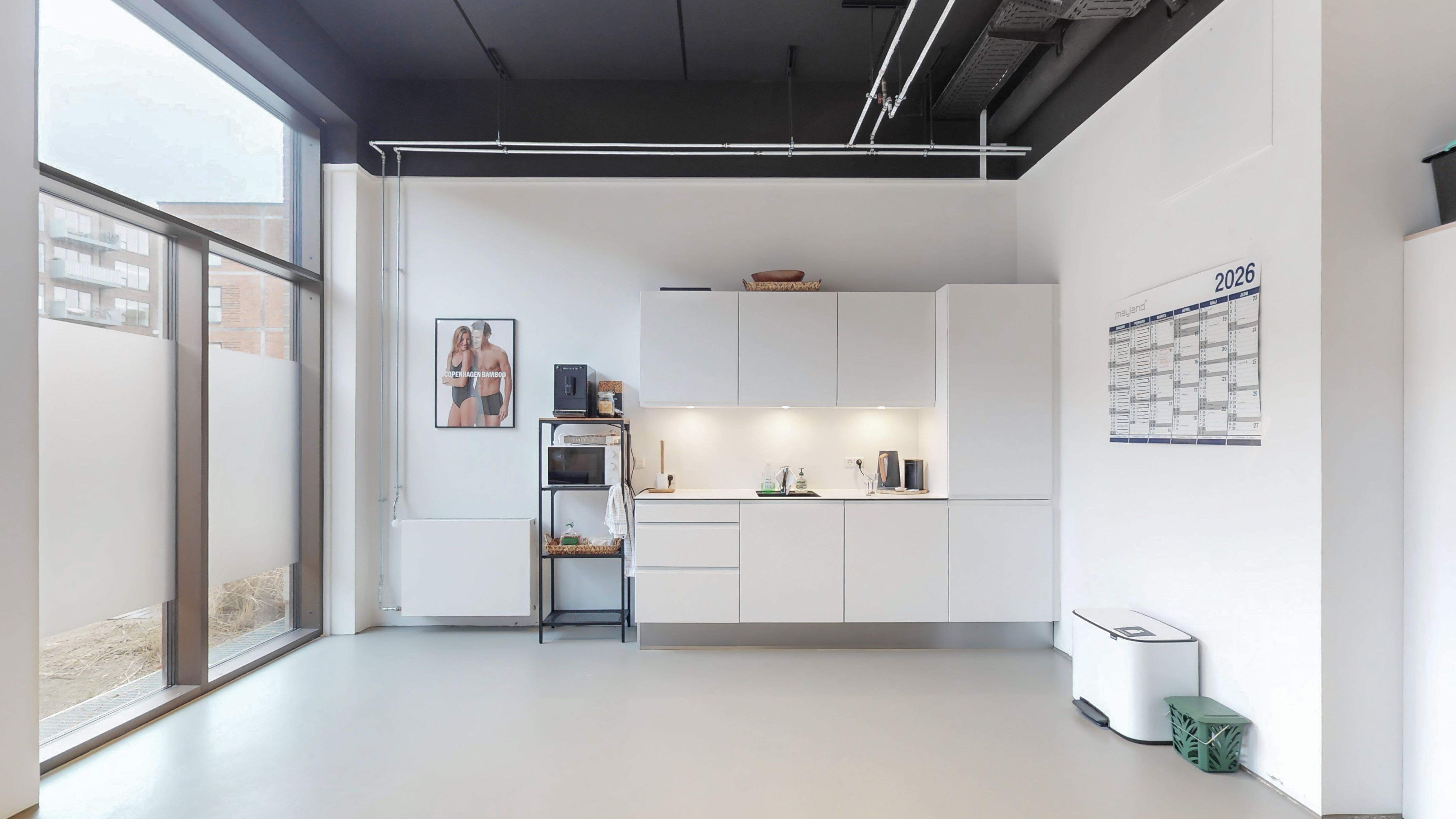 Lyst og funktionelt kontorlokale med hvide vægge og loft. En stor vinduesglas og gulv. En kalender og et billede hænger på væggen. Et skab med et mikroovn og kaffemaskine er placeret ved væggen.