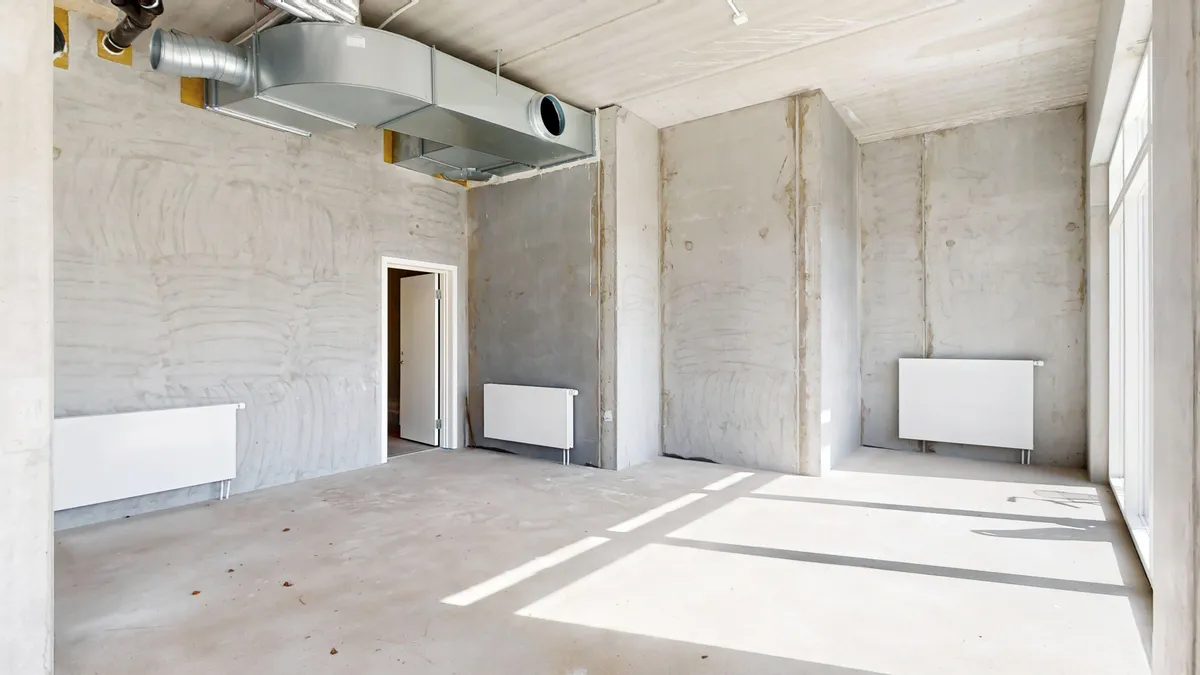 En stor tom lejlighed med hvid gulv og vægge samt to lyse radiatorer og en ventil i loftet