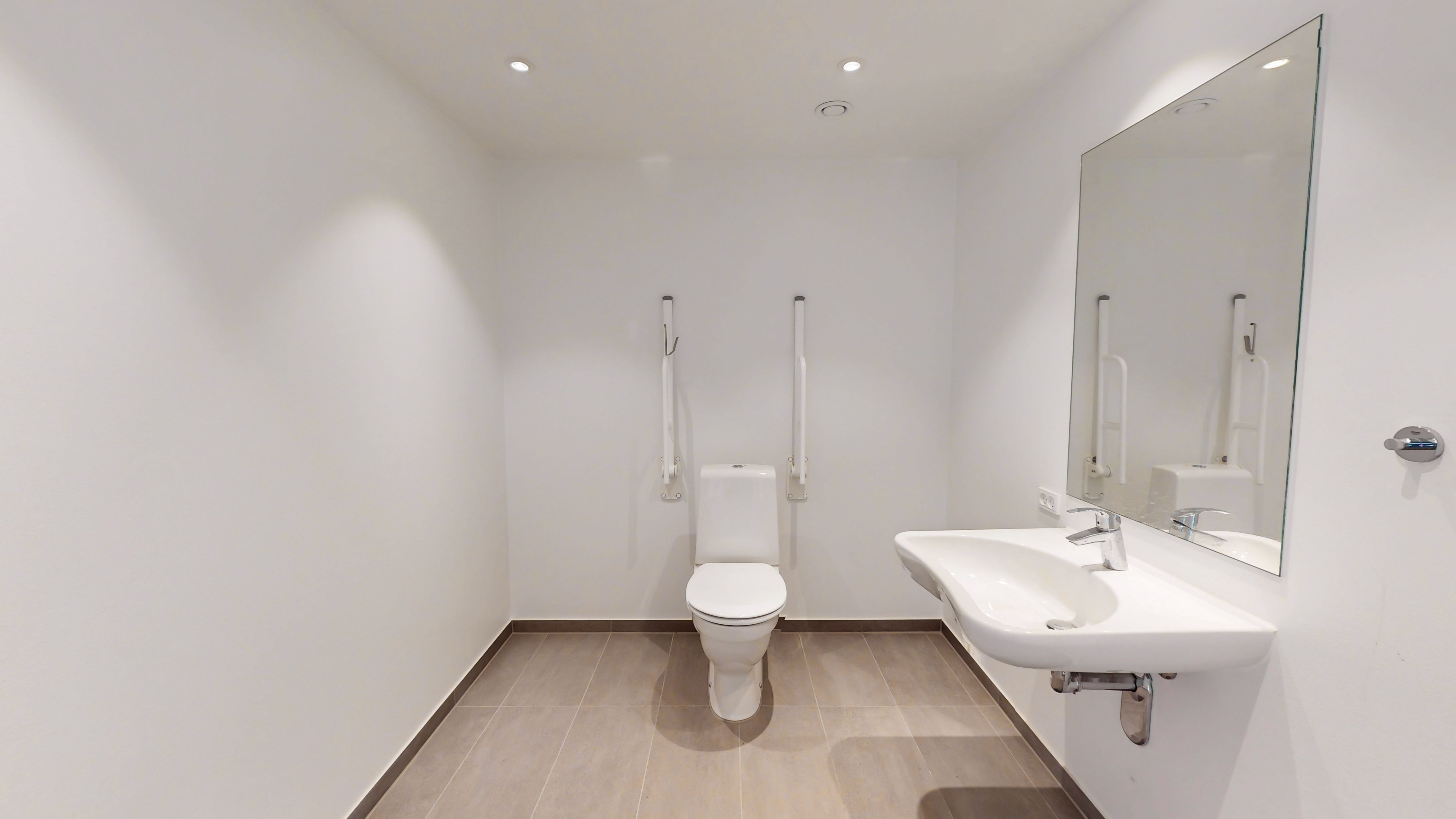 Alt-tekst: Rengøringsvenligt hjørnelejemål med toilet, håndvask og spejl i hvid interiør med gulvbeklædning og lette gulvprofiler