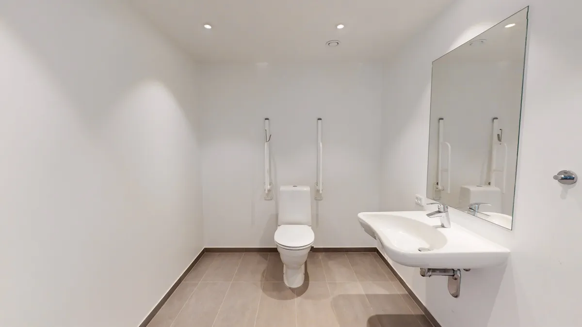 Alt-tekst: Rengøringsvenligt hjørnelejemål med toilet, håndvask og spejl i hvid interiør med gulvbeklædning og lette gulvprofiler