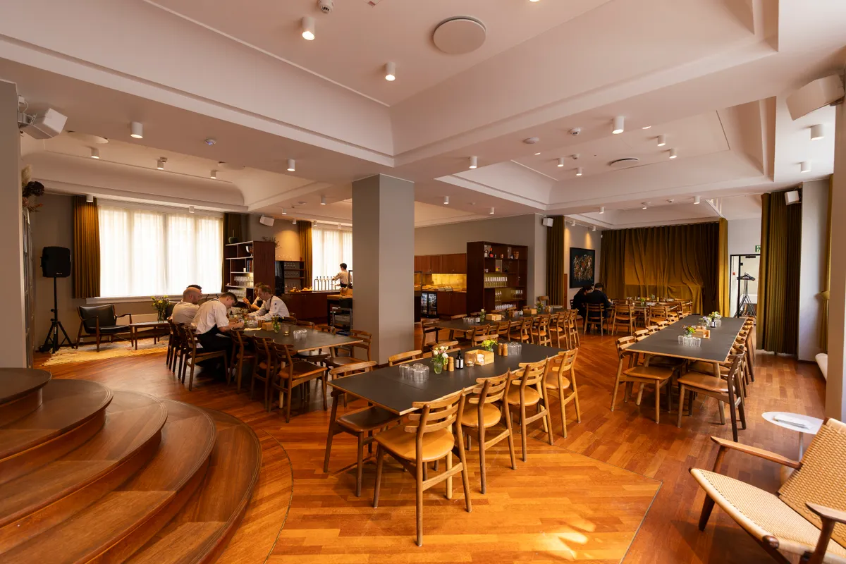 Lejlighed med møbler og dekorationer. Mennesker sidder og drikker ved lange borde. Køkken med øl i køleskab og serveringsbord. Gulvtæppe og højt loft med indbygget belysning.