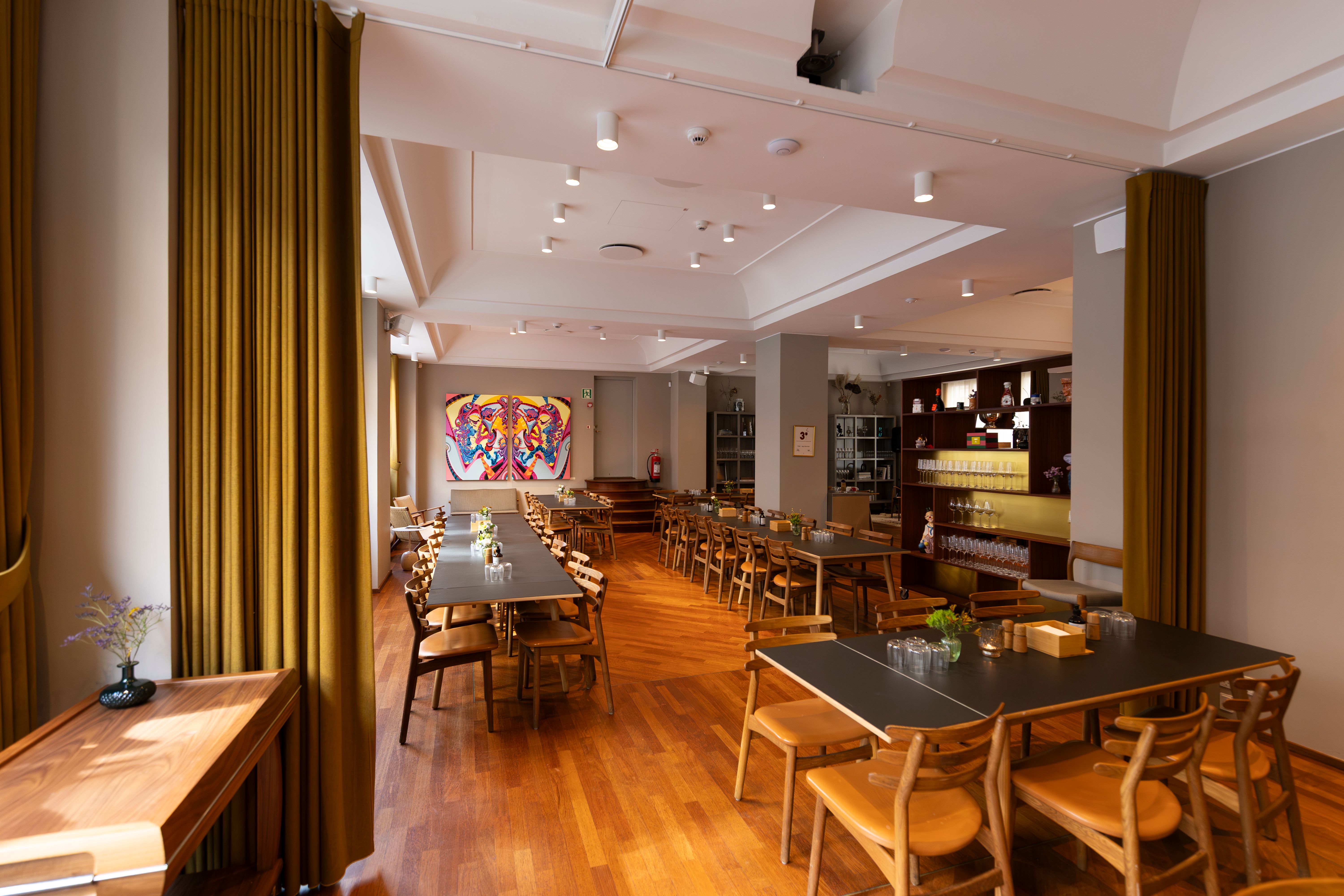 Lys, elegant mødelokale med lysegul væg og mønstret gulv, to lange borde og stole, skrå loft og mønstret gulv, hylder og kunstværker på baggrunden