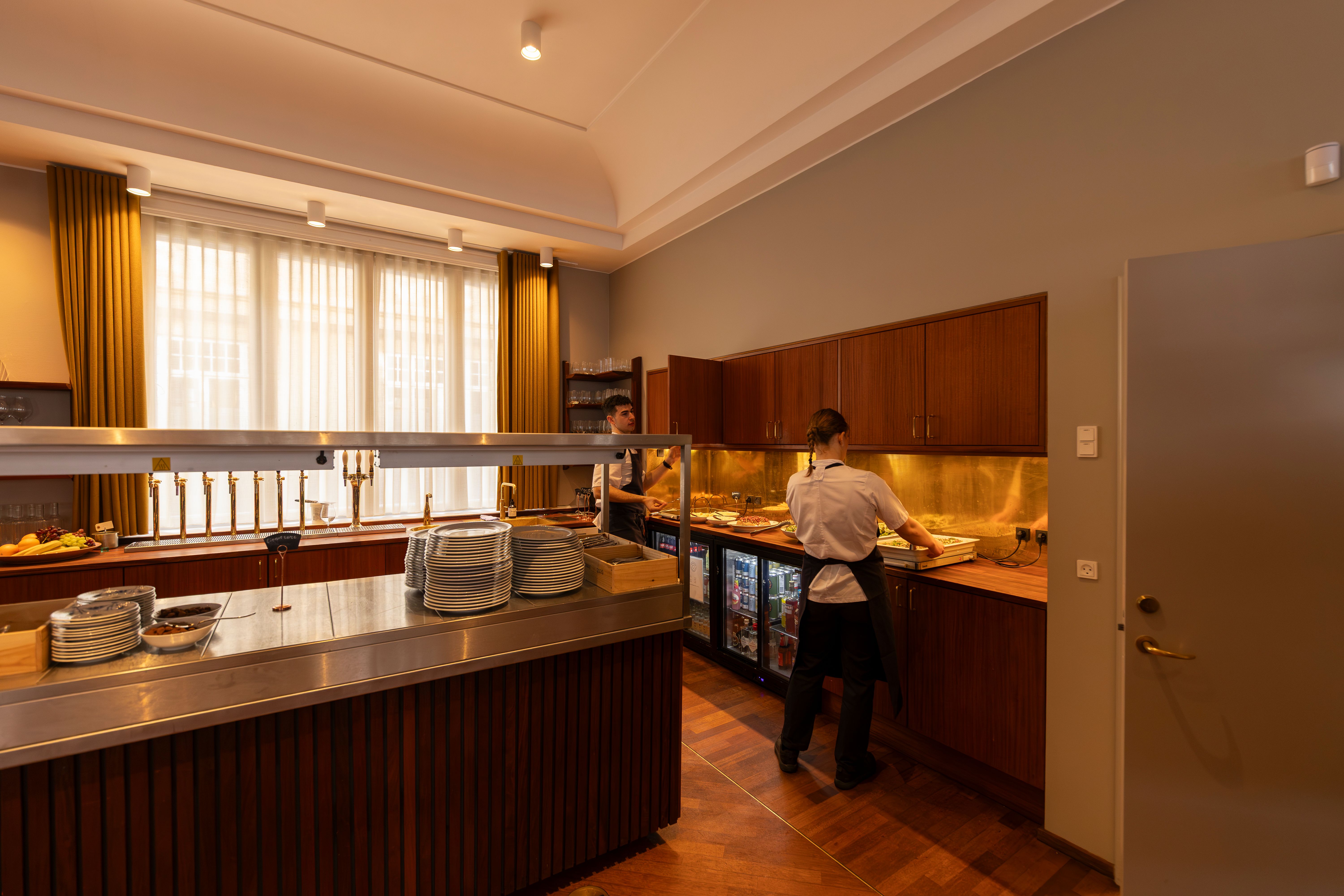 To medarbejdere i en lukket café med stolpeglas og lysekroner, der serverer gæster i en luksuriøs restaurant med lysegulle vægge og træindretning