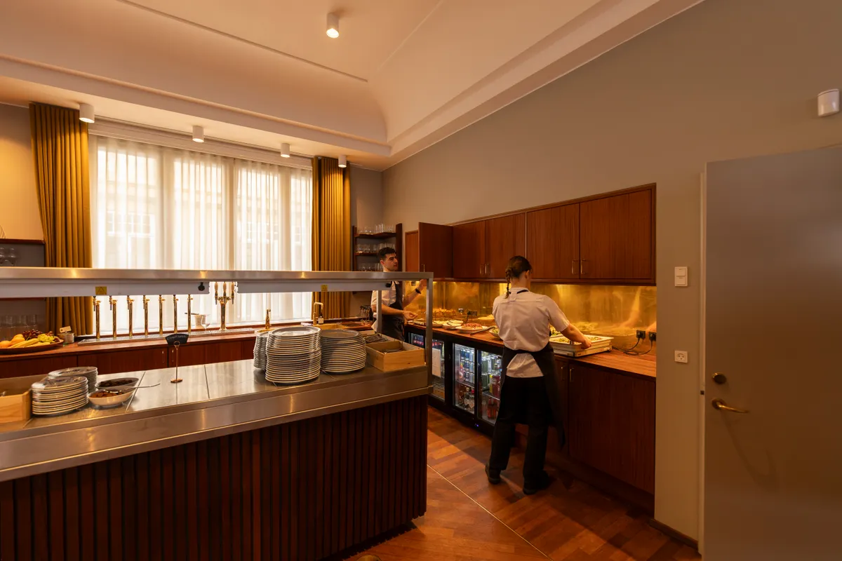 To medarbejdere i en lukket café med stolpeglas og lysekroner, der serverer gæster i en luksuriøs restaurant med lysegulle vægge og træindretning