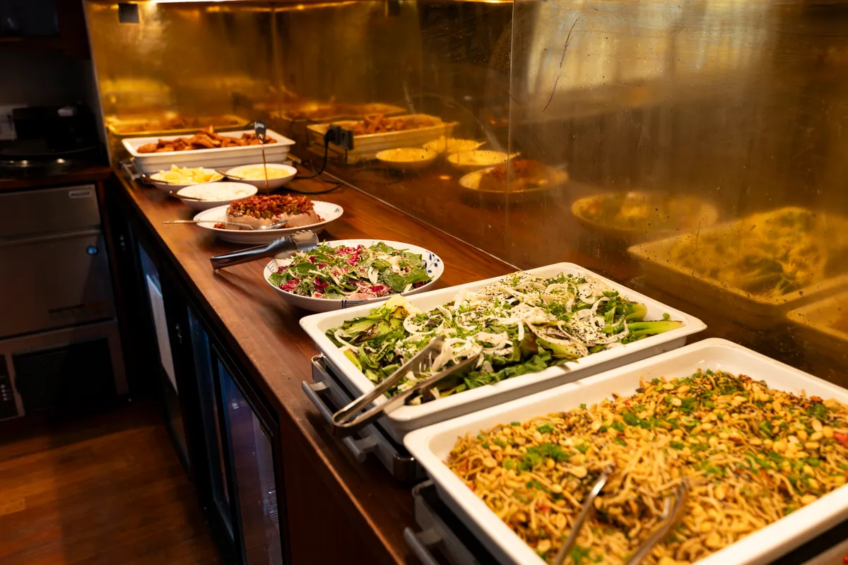 Buffet med flere skåle og tallerkener med grøntsager og salat på et bord med træoverflade, der er placeret i et kontormiljø