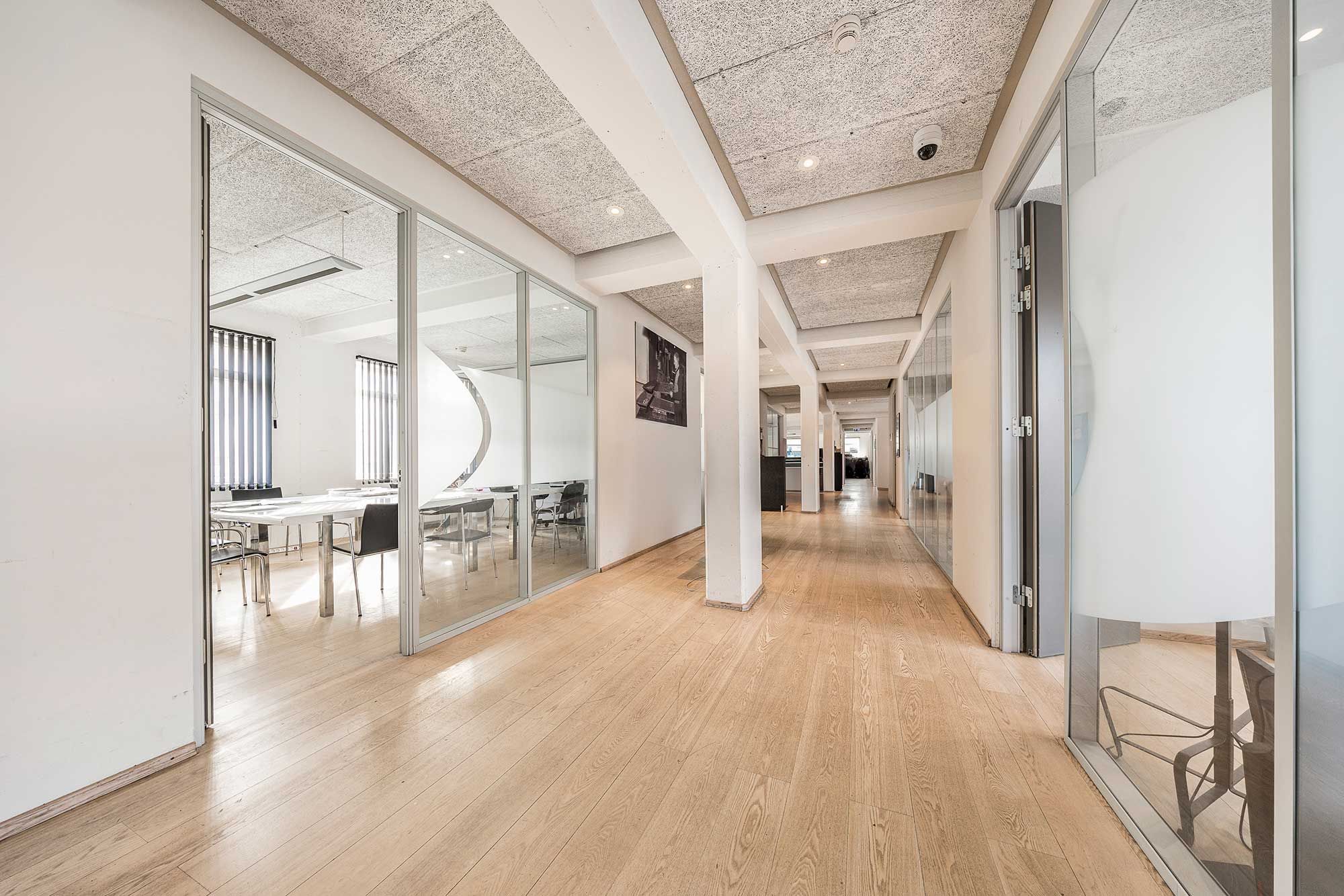 Lyst kontor med hvide vægge, store glasruder, lyse lofter, lysegrå loftspaneler, hårdt trægulv, møbler og borde, åbne døre og slanke søjler