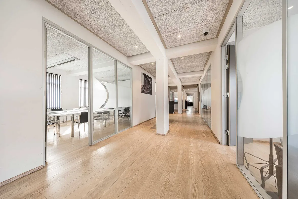 Lyst kontor med hvide vægge, store glasruder, lyse lofter, lysegrå loftspaneler, hårdt trægulv, møbler og borde, åbne døre og slanke søjler