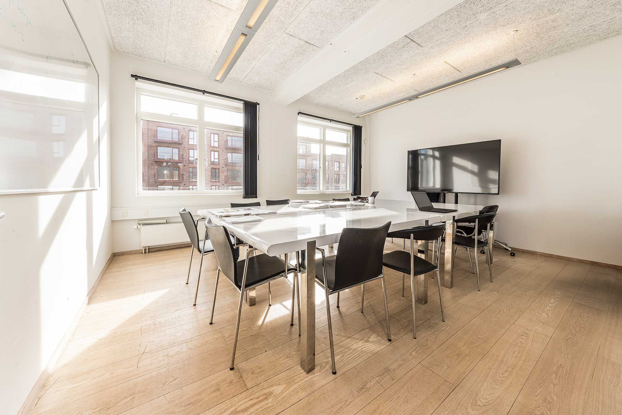 Kontorlokaler med hvidpudsede vægge, moderne møbler og en stor, hvid bord med flere stole. Vindue med sort gardin og en stor, flad skærm i væggen.