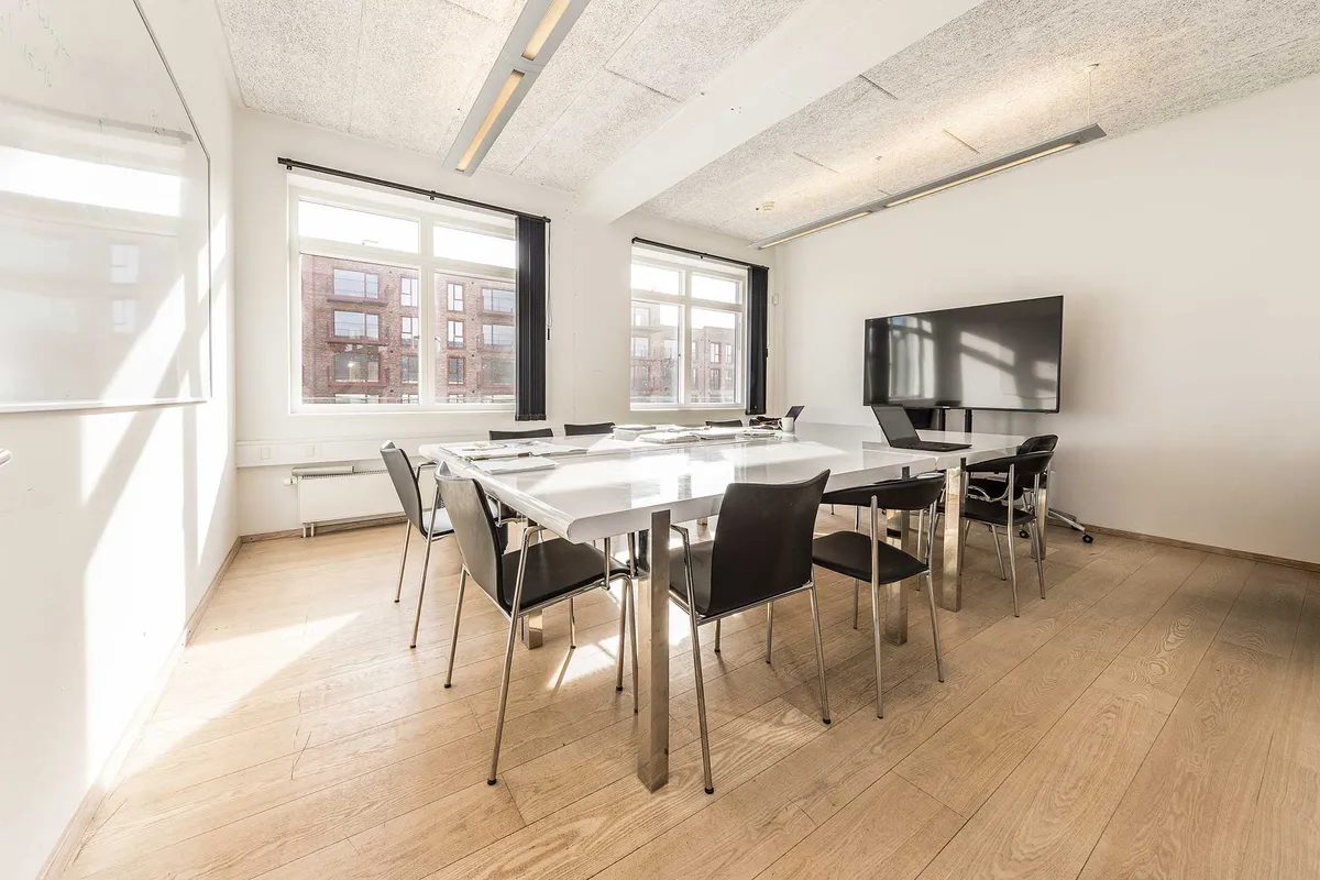 Kontorlokaler med hvidpudsede vægge, moderne møbler og en stor, hvid bord med flere stole. Vindue med sort gardin og en stor, flad skærm i væggen.