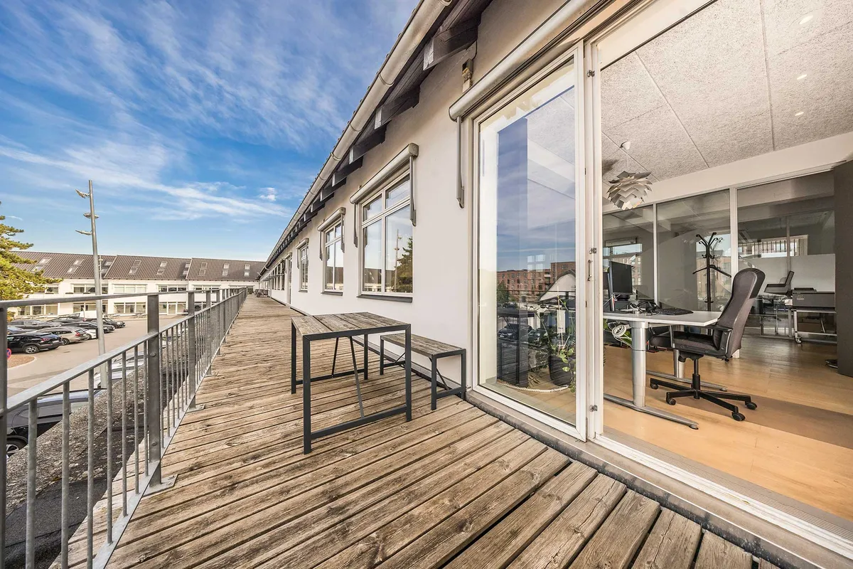 Kontorloft med trægulv og metalgeværer med udsigt over parkeringsplads og bygninger med hvide facader og tegltag.