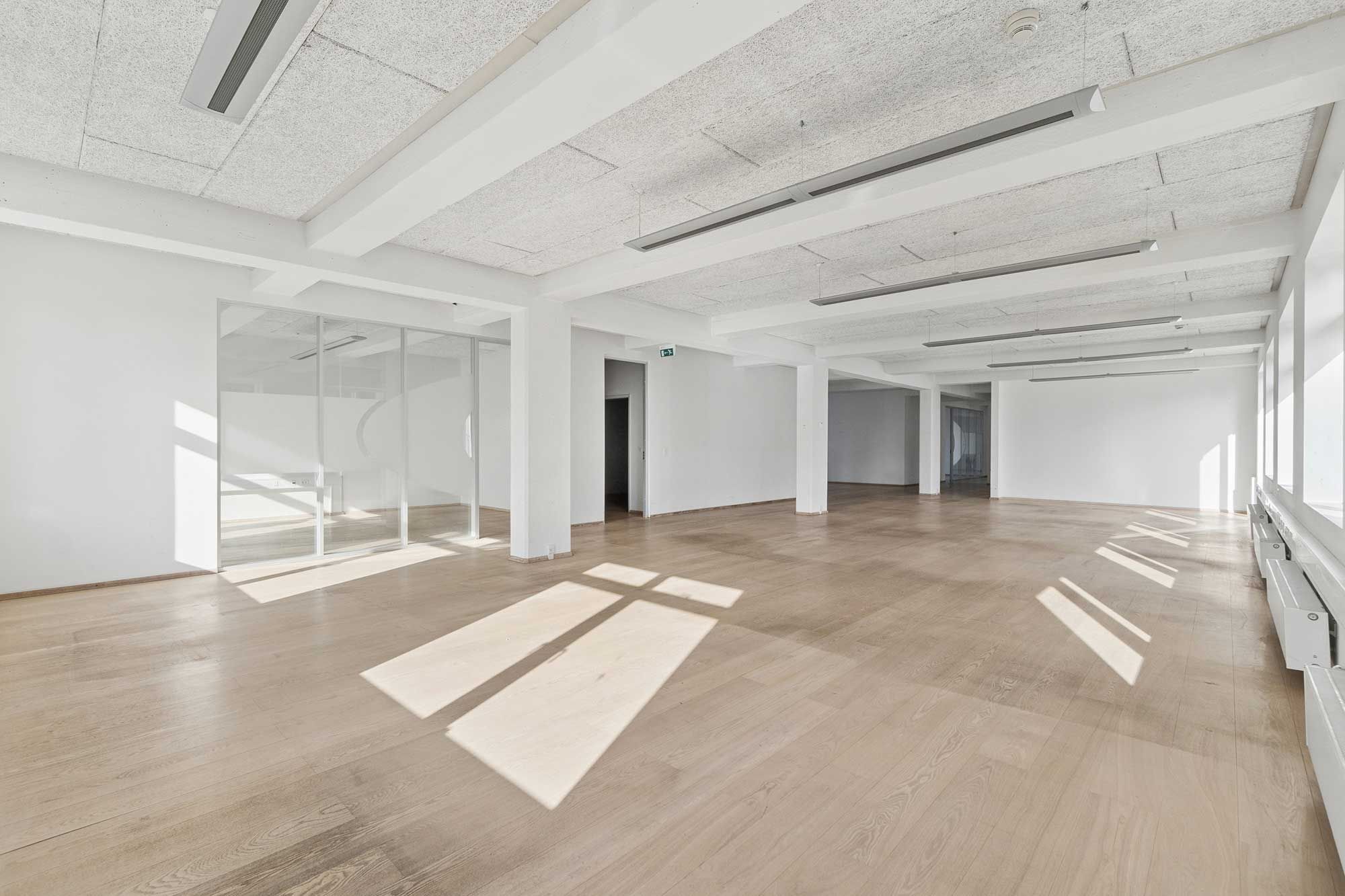 Et lyst kontorlokal med hvide vægge og lyse trægulve. Det er et meget stort lokale med store glasruder og højt til loftet. Der er lyse stråler af solen på gulvet