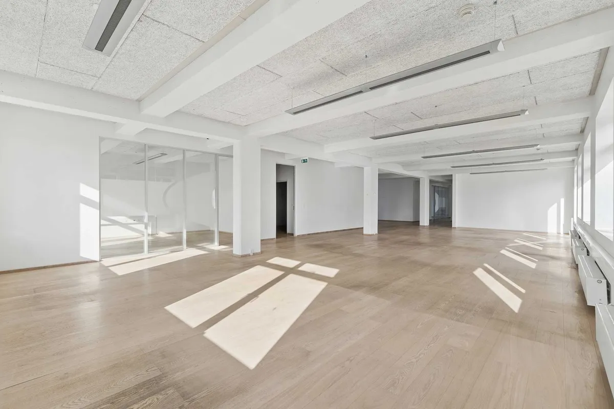 Et lyst kontorlokal med hvide vægge og lyse trægulve. Det er et meget stort lokale med store glasruder og højt til loftet. Der er lyse stråler af solen på gulvet