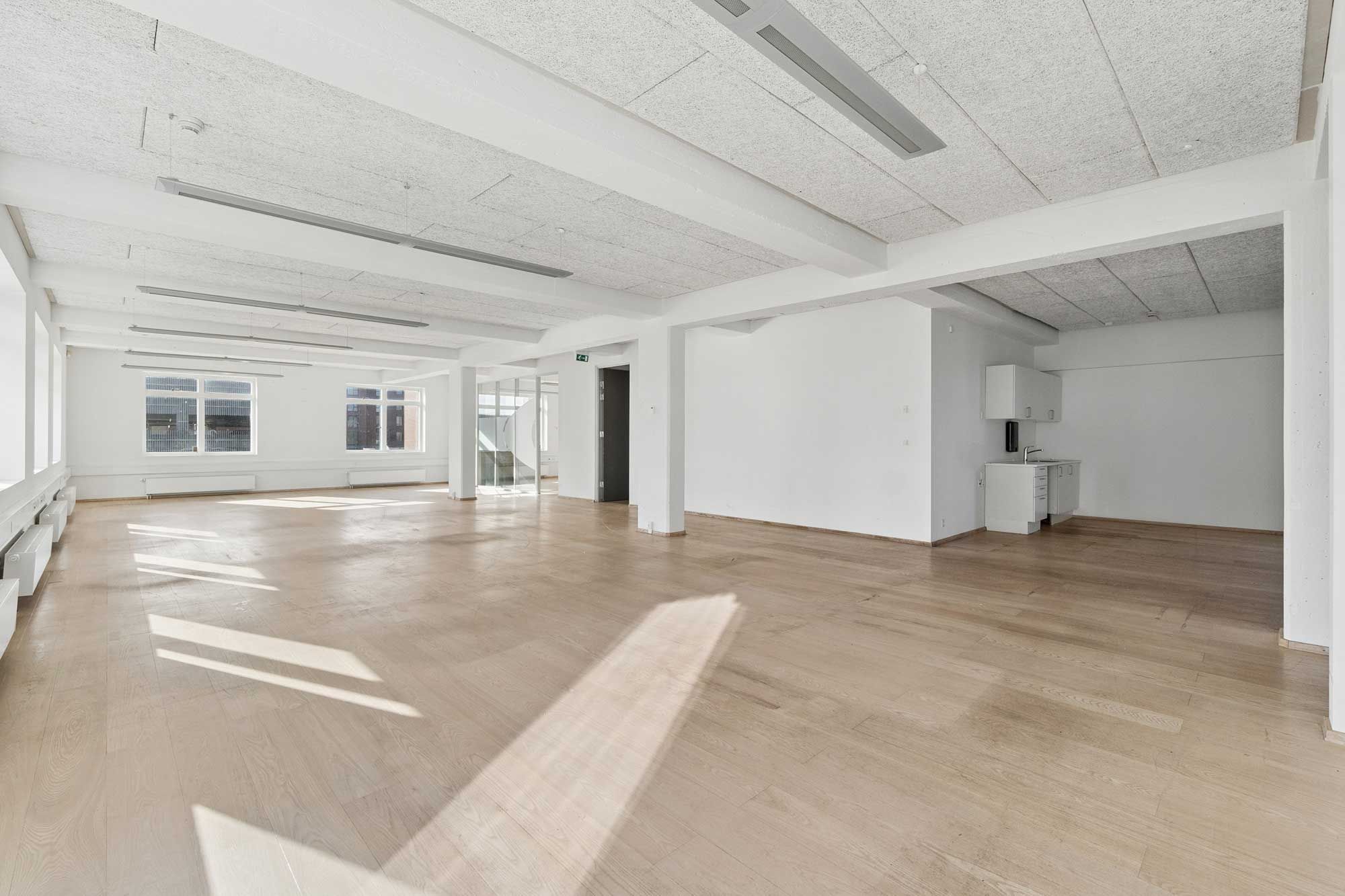 En stor, tom kontorlokation med hvide vægge, højt til loftet, lyse loftsbjælker og lyse gulve med en lille køkkendel og vinduer med udsigt til gården