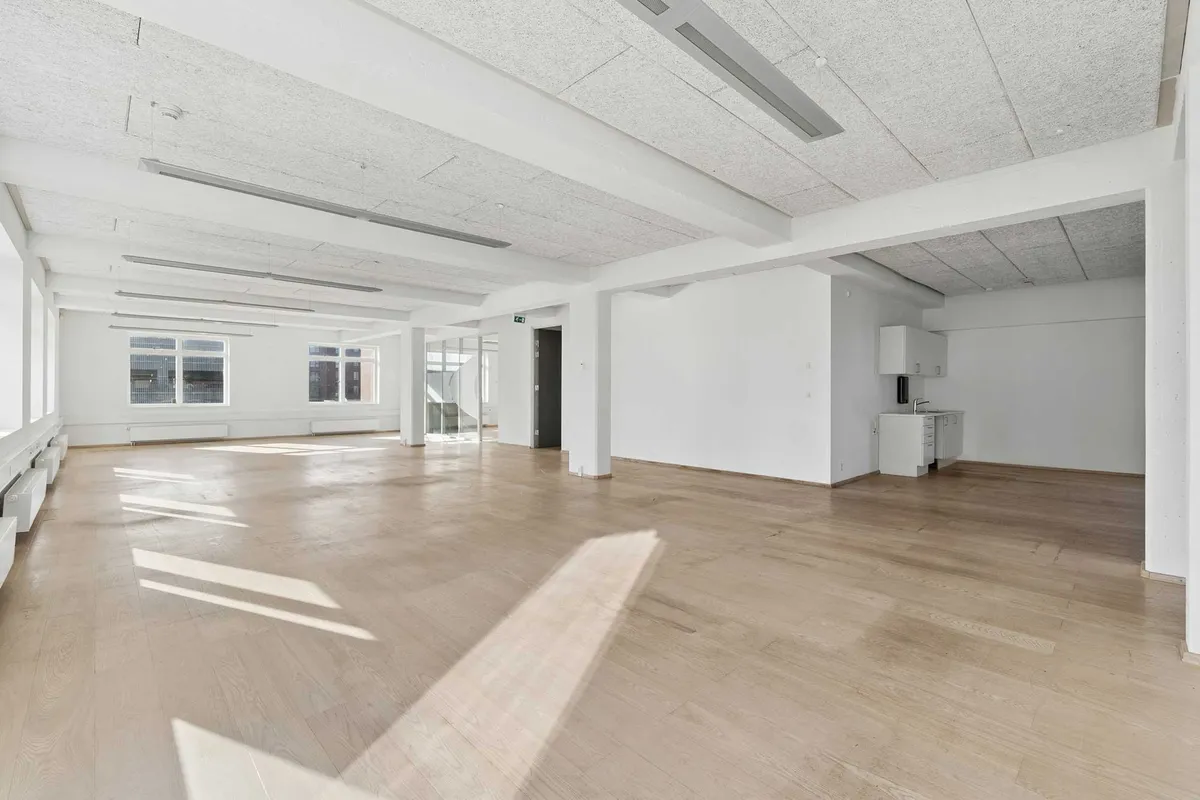 En stor, tom kontorlokation med hvide vægge, højt til loftet, lyse loftsbjælker og lyse gulve med en lille køkkendel og vinduer med udsigt til gården