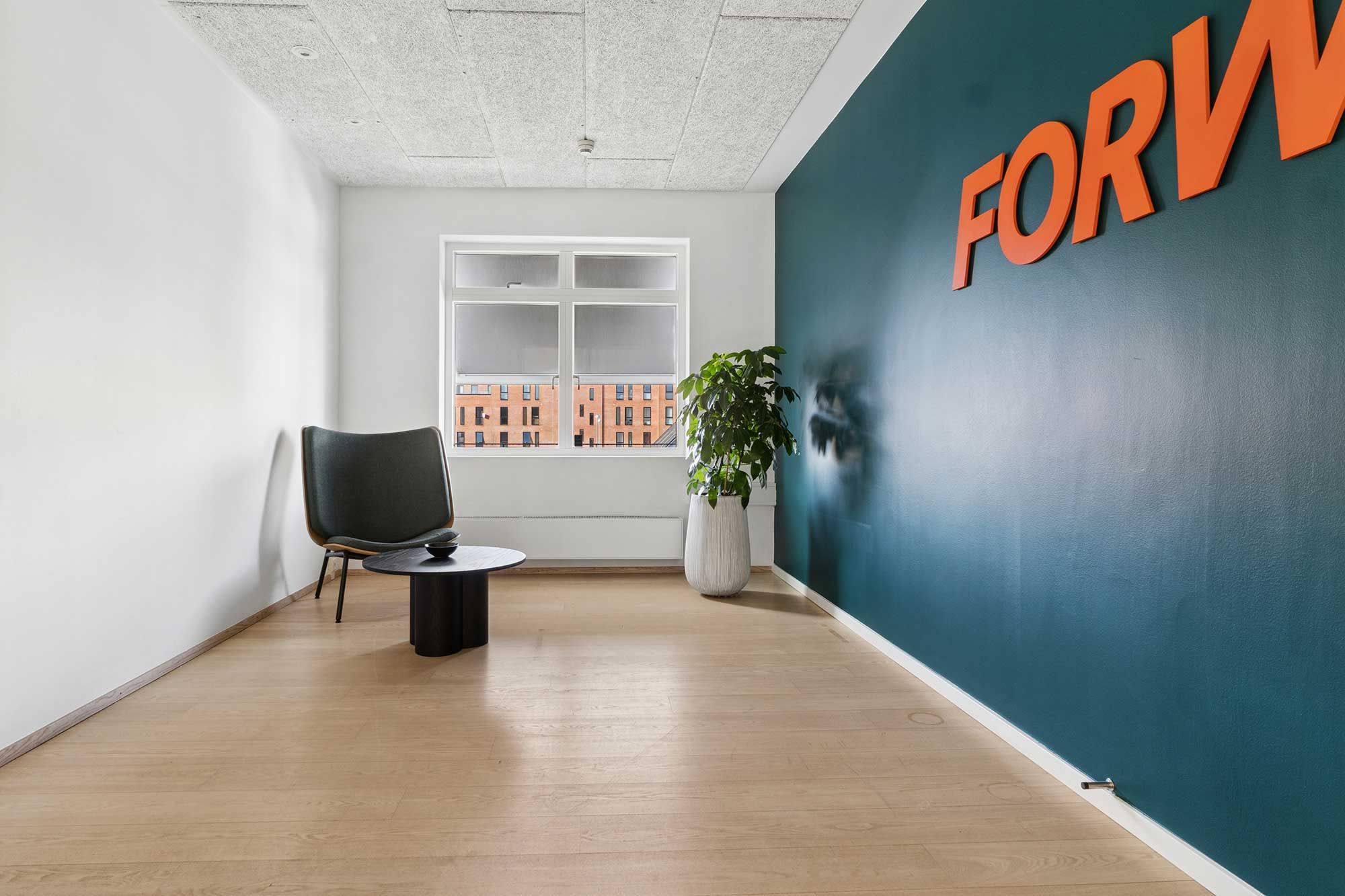 Et lyse og rummeligt kontor med højloft og pænt gulv, møbel med plante og træbord, samt lysevinduer med udsigt til bygninger