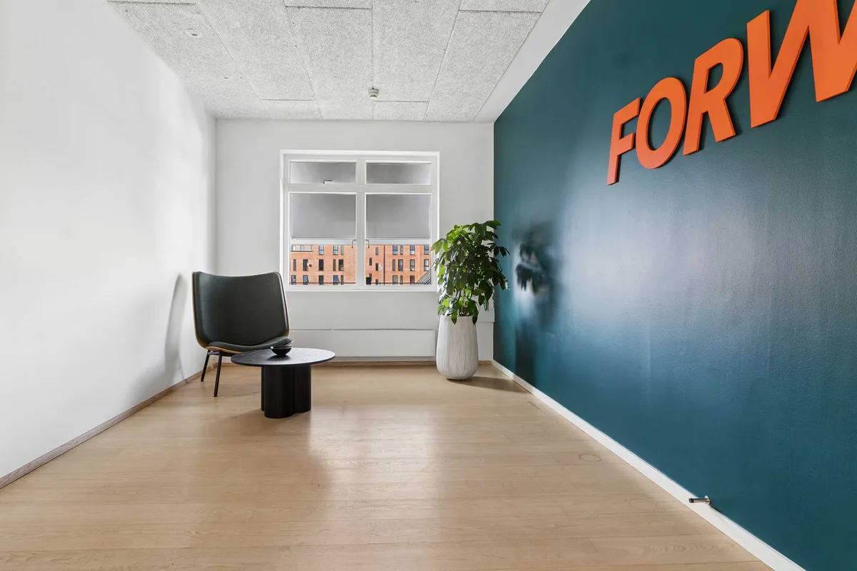 Et lyse og rummeligt kontor med højloft og pænt gulv, møbel med plante og træbord, samt lysevinduer med udsigt til bygninger