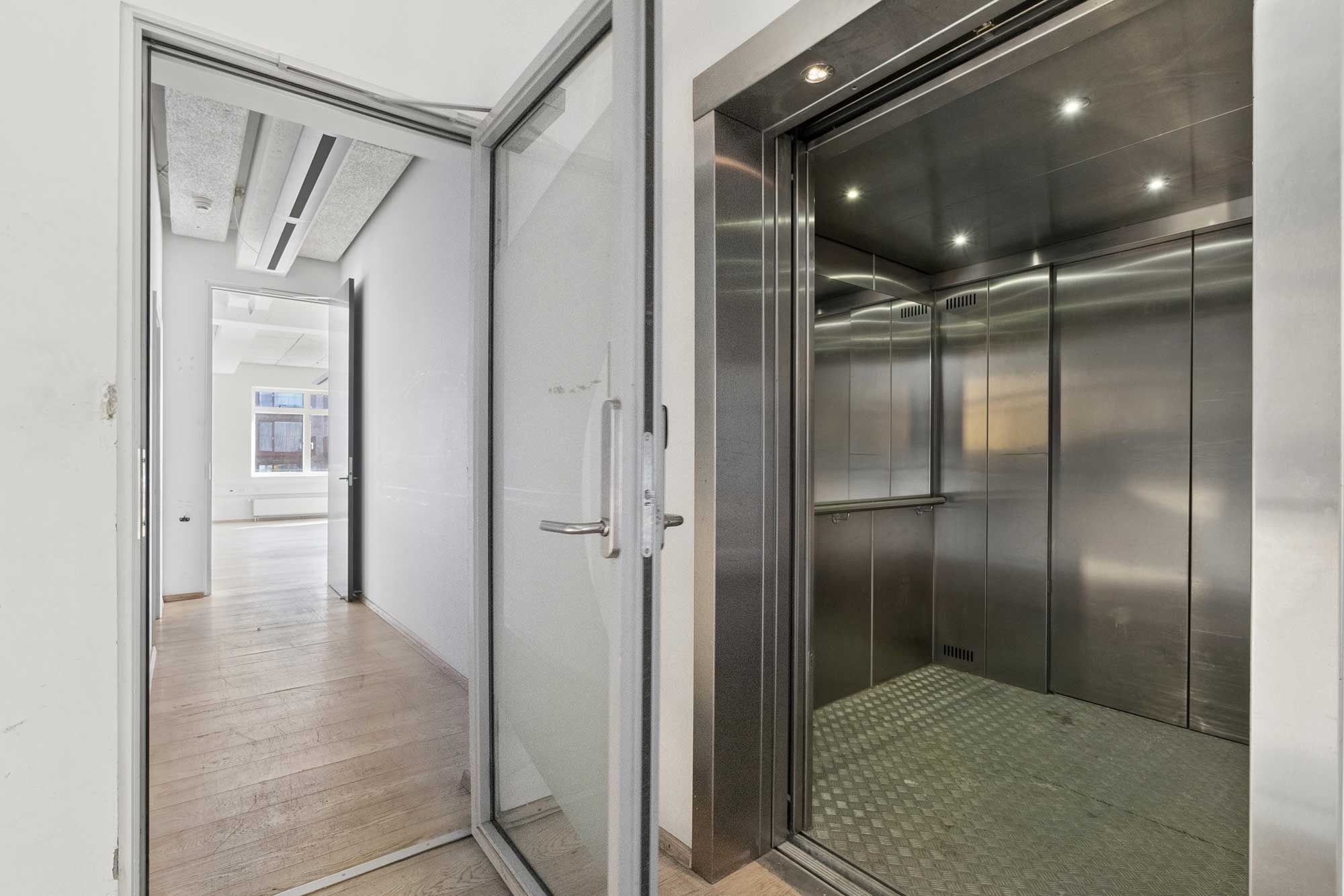 I fotoet ses en åben elevatordør med en blank gulvbelægning og hvide vægge. Der er lyskaster på loftet. Der er en åben dør i baggrunden med en udsigt til en kantine