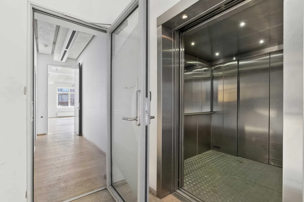 I fotoet ses en åben elevatordør med en blank gulvbelægning og hvide vægge. Der er lyskaster på loftet. Der er en åben dør i baggrunden med en udsigt til en kantine