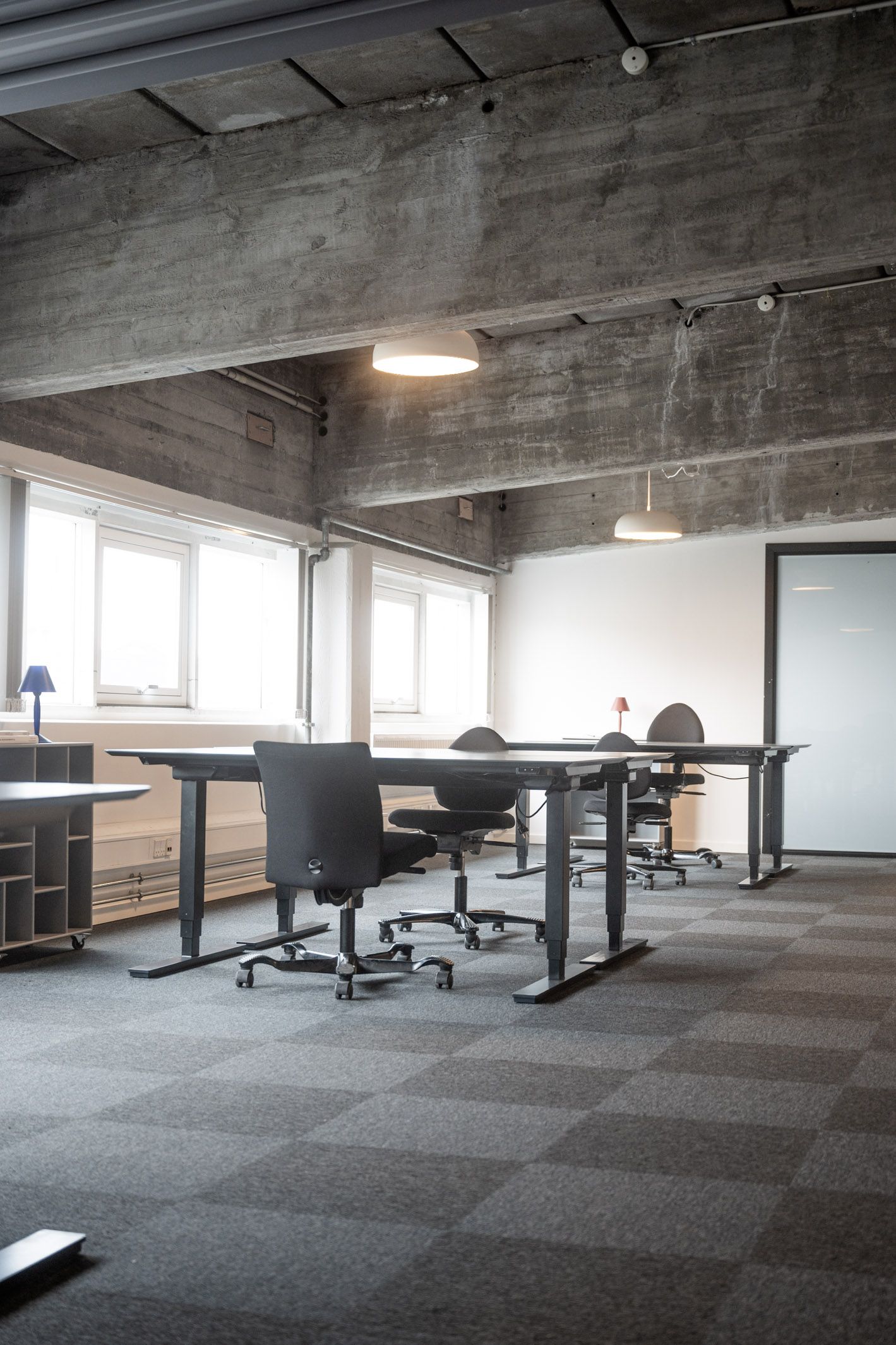 Række med stol og bord i et kontor med højt loft og lyse vindue. Gulvet er lysegrå og der er en hvid dør i baggrunden