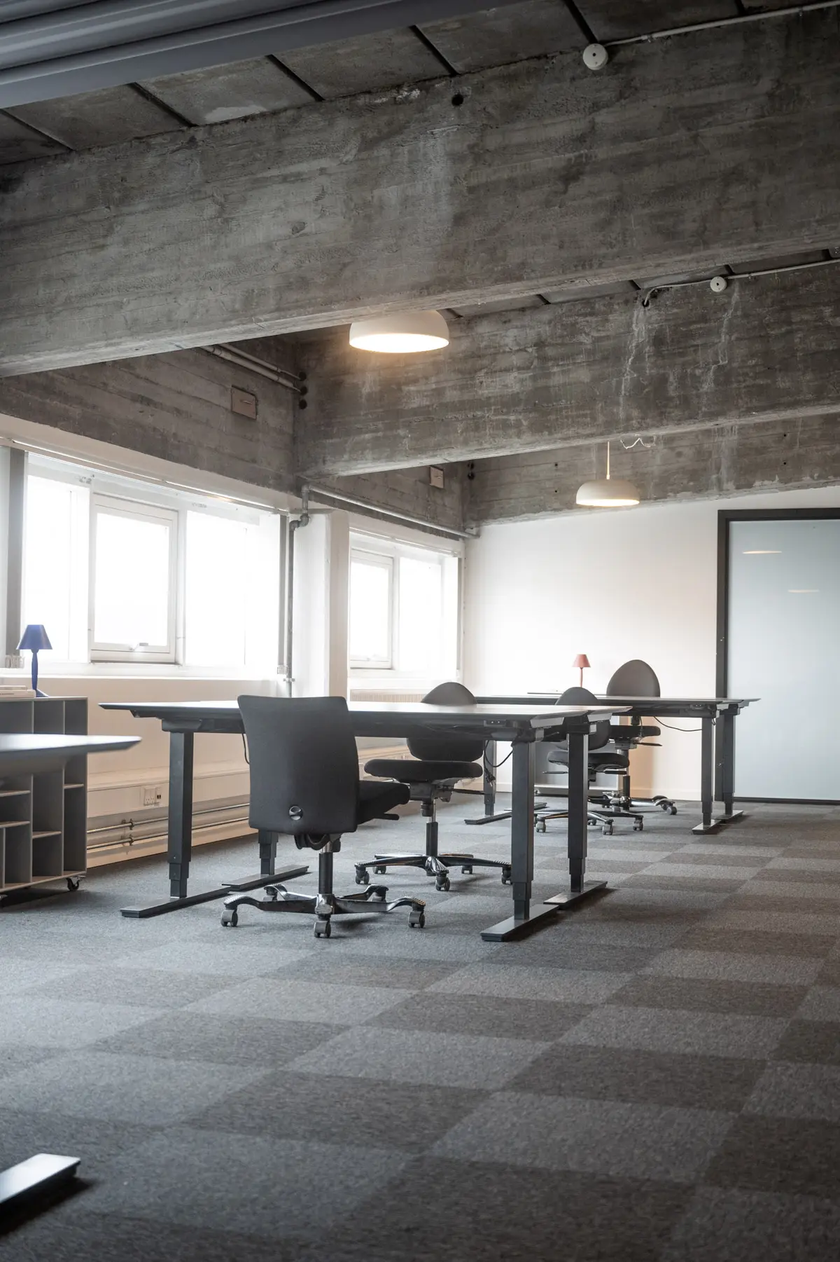 Række med stol og bord i et kontor med højt loft og lyse vindue. Gulvet er lysegrå og der er en hvid dør i baggrunden