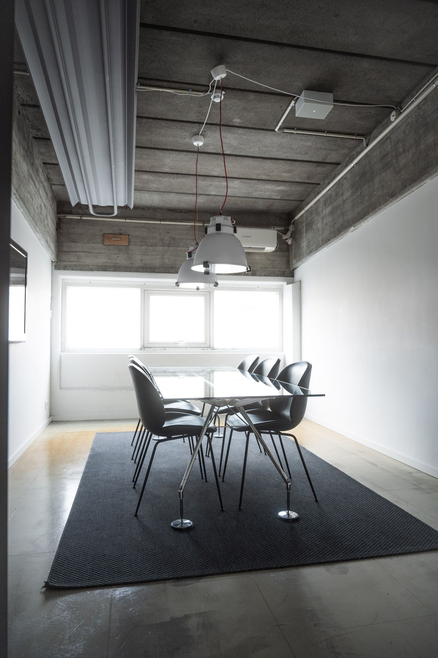 Kontor med gulvbelægning, vinduer, to hængelampe og glasbord samt stol. I loftet er der flere røre og lamper.