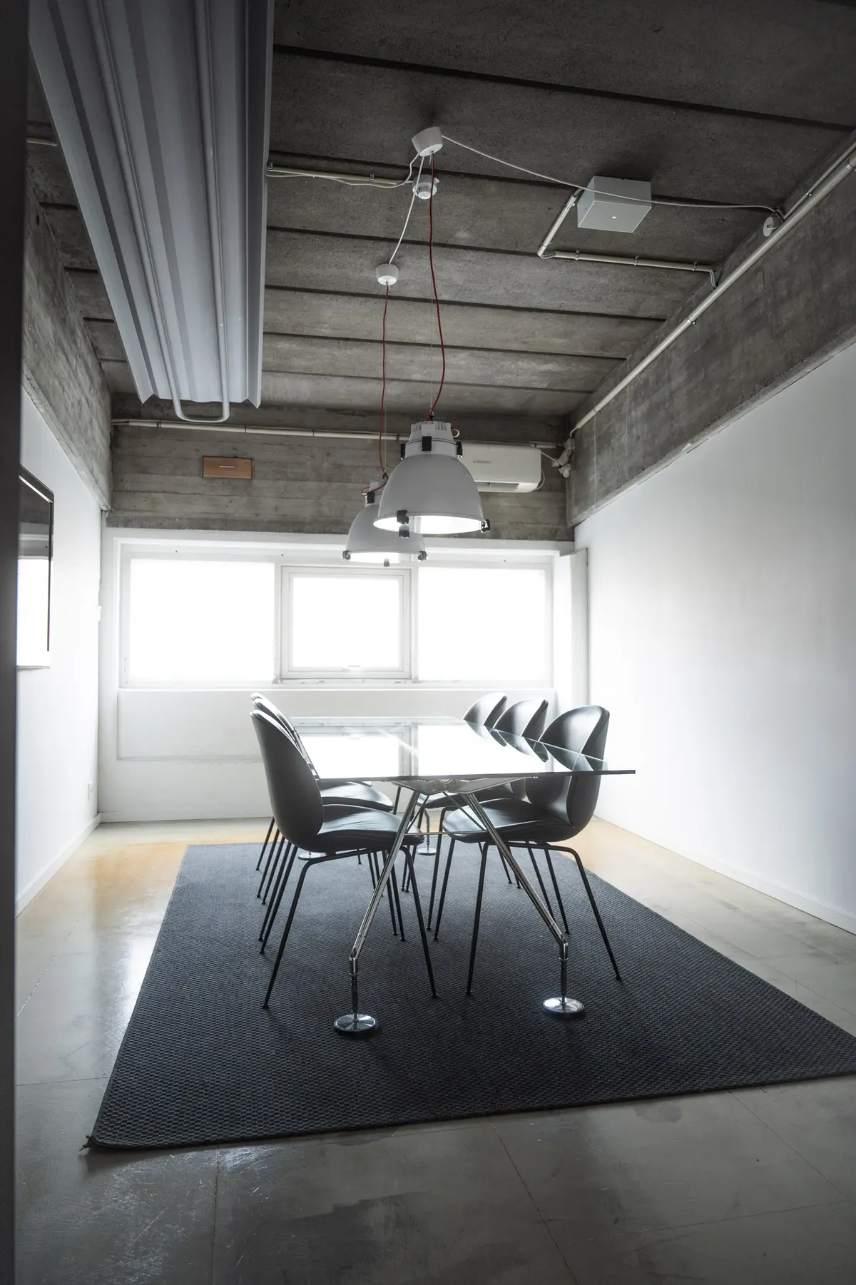 Kontor med gulvbelægning, vinduer, to hængelampe og glasbord samt stol. I loftet er der flere røre og lamper.