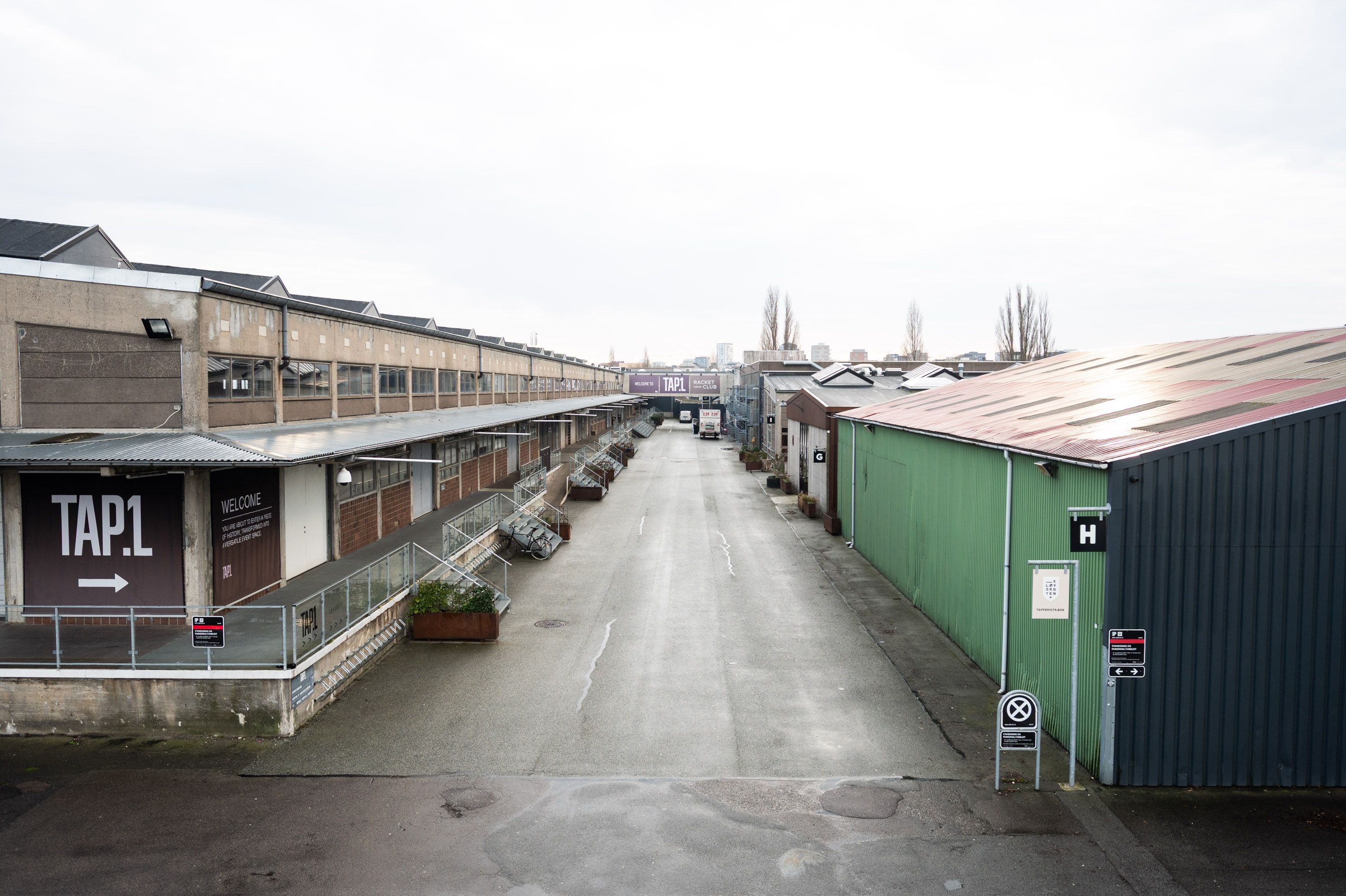 Lang vej med gamle lagerbygninger og skilte med tekst og pile, der fører til en lukket dør og flere bygninger med skilte