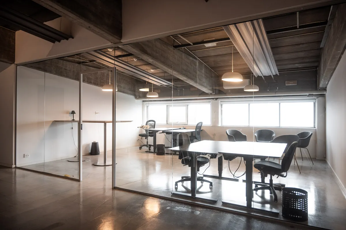To bord, to stole og en lampestativ i et kontor med høje ruder og loftsbjælker, der er delvist synlige gennem glasvægge. Konferencebordene er lyse og minimalistiske med høje stolpe