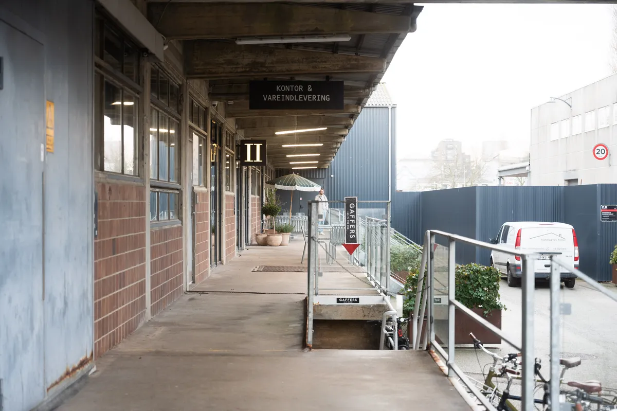 To kontorlokaler med glasdøre og trægulv, hvor der er skilte med tekst og tal samt en hvid varevogn parkeret og to cykler stående til højre