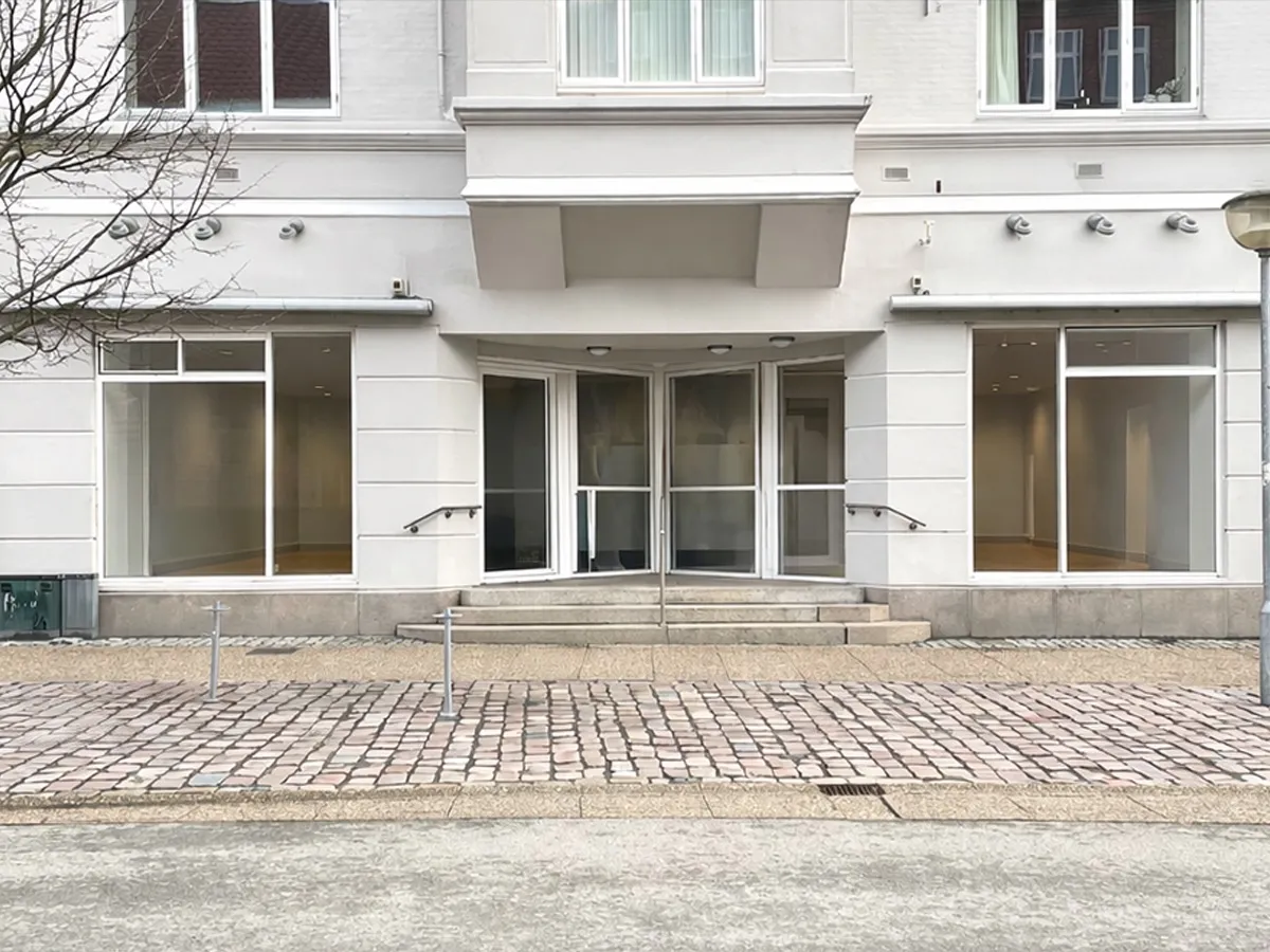 Lejlighed til leje på Østergade 11 i Hjørring med stort vindue og høj synlighed. Indgang med skrappe trin og to håndsvæve, der fører ind i bygningen.