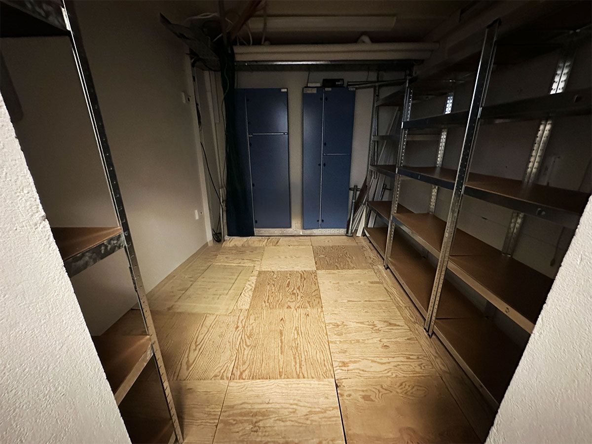 Lager med nyt trægulv og lagerhylder, hvor loftet er højt og der er to blå skabe. Gulvet er let lysebrunt og trægulvet er lige nylagt.