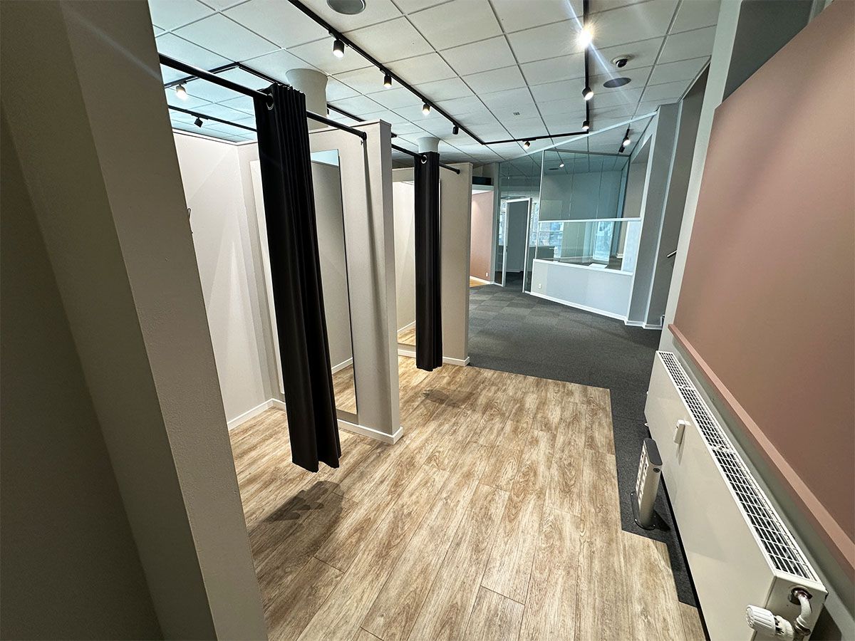 To store lokaler med hvide vægge og gulv samt højt loft med lys. To lokaler er adskilt med sorte gardiner og der er gulvtæppe på gulvet.