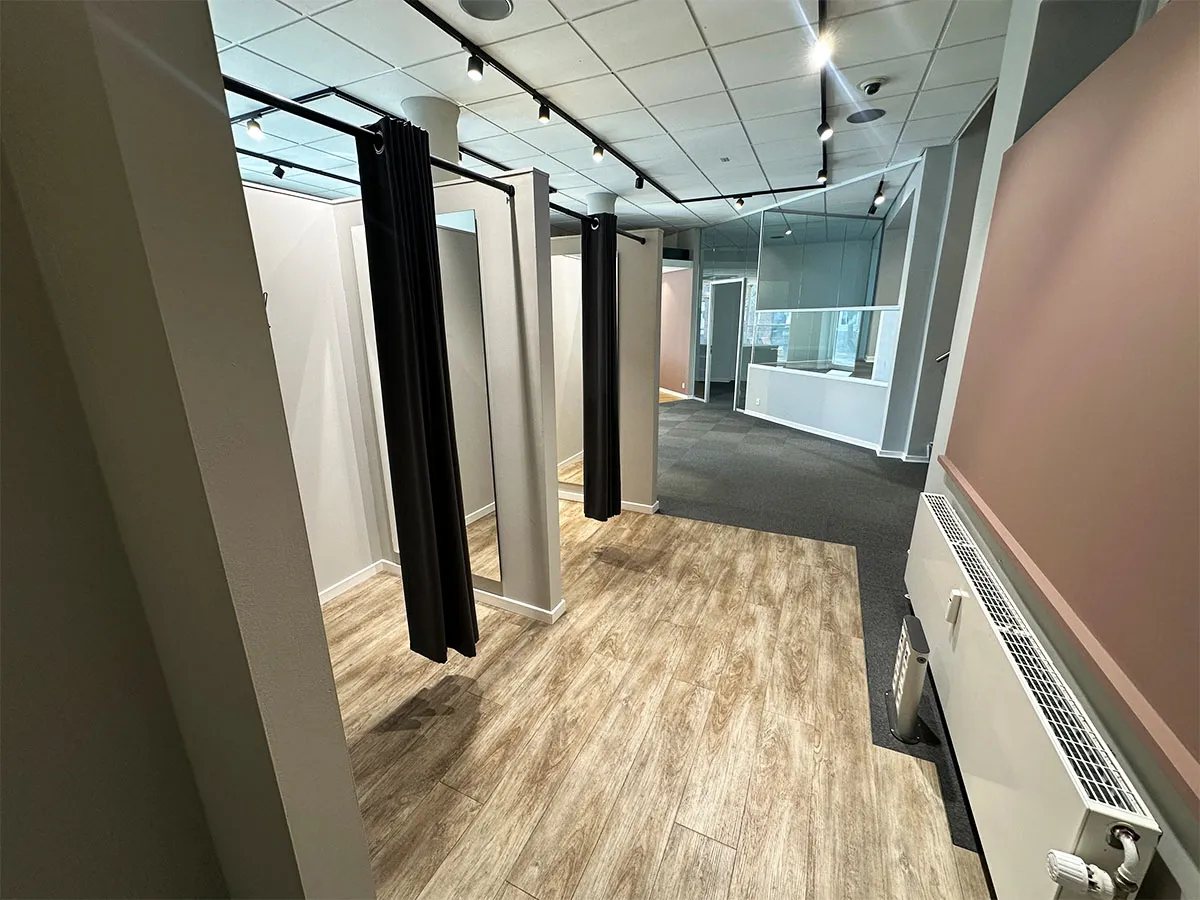 To store lokaler med hvide vægge og gulv samt højt loft med lys. To lokaler er adskilt med sorte gardiner og der er gulvtæppe på gulvet.