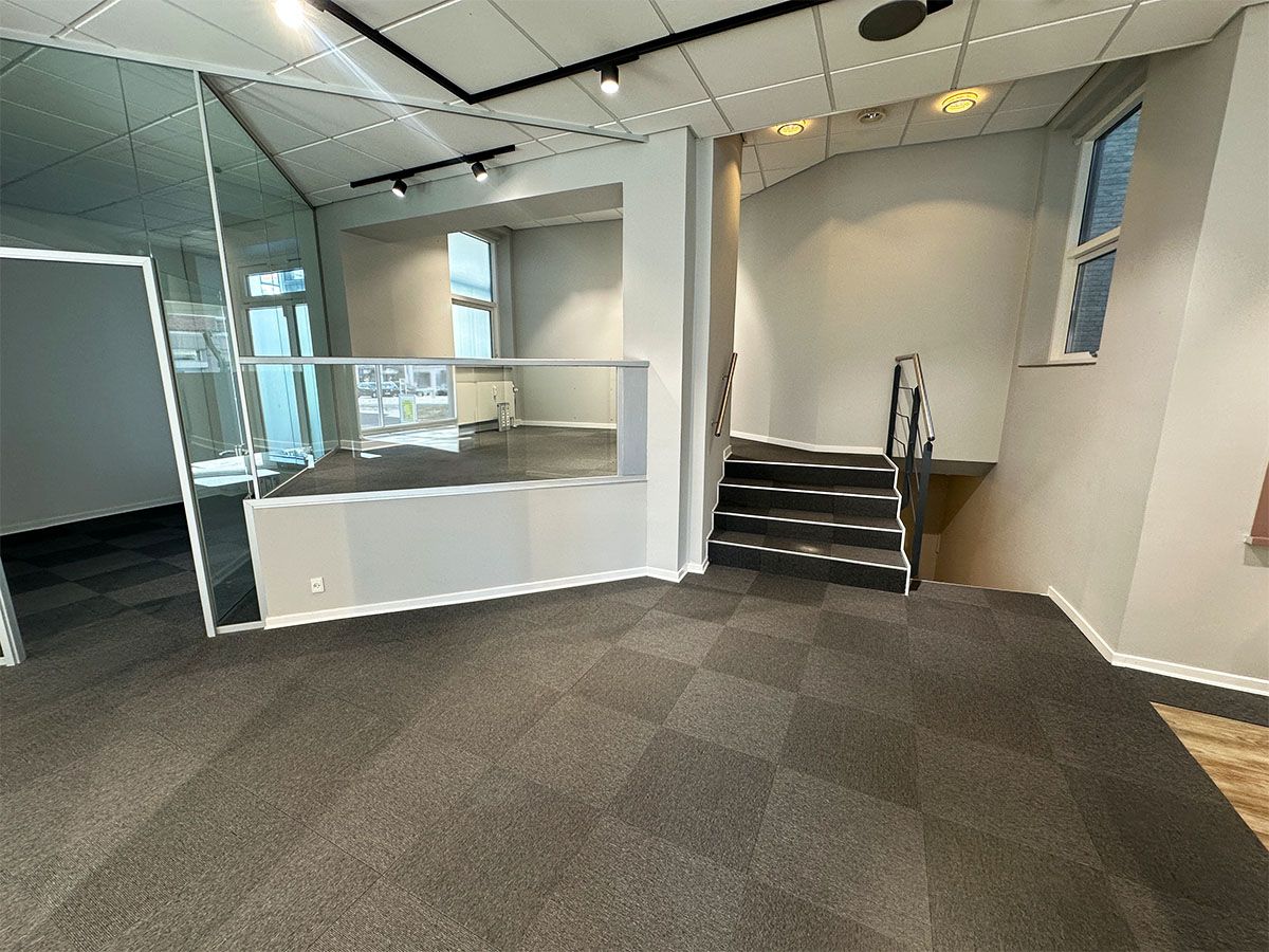 Store lokale med glasdør og trapper til øverste etage. Hvid og grå farvelægning. Moderne indretning med krydsformede loftslys og gulvbelægning.